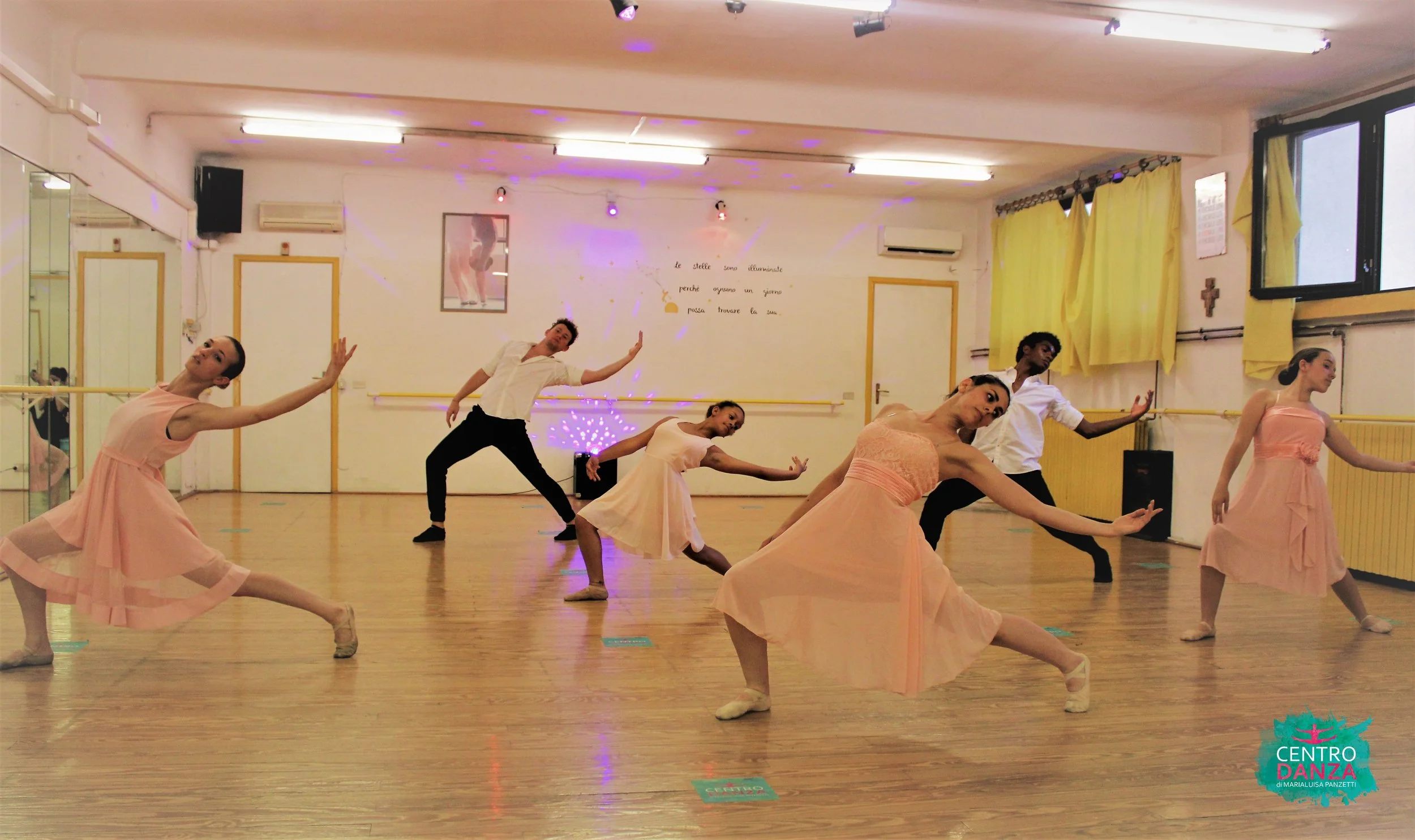 Ballerini in un balletto di gruppo in una sala di danza. Le ballerine indossano lunghi tutù rosa, mentre i ballerini indossano pantaloni neri e camicia bianca. La scena è illuminata con luci colorate e sul muro c'è una frase in italiano.