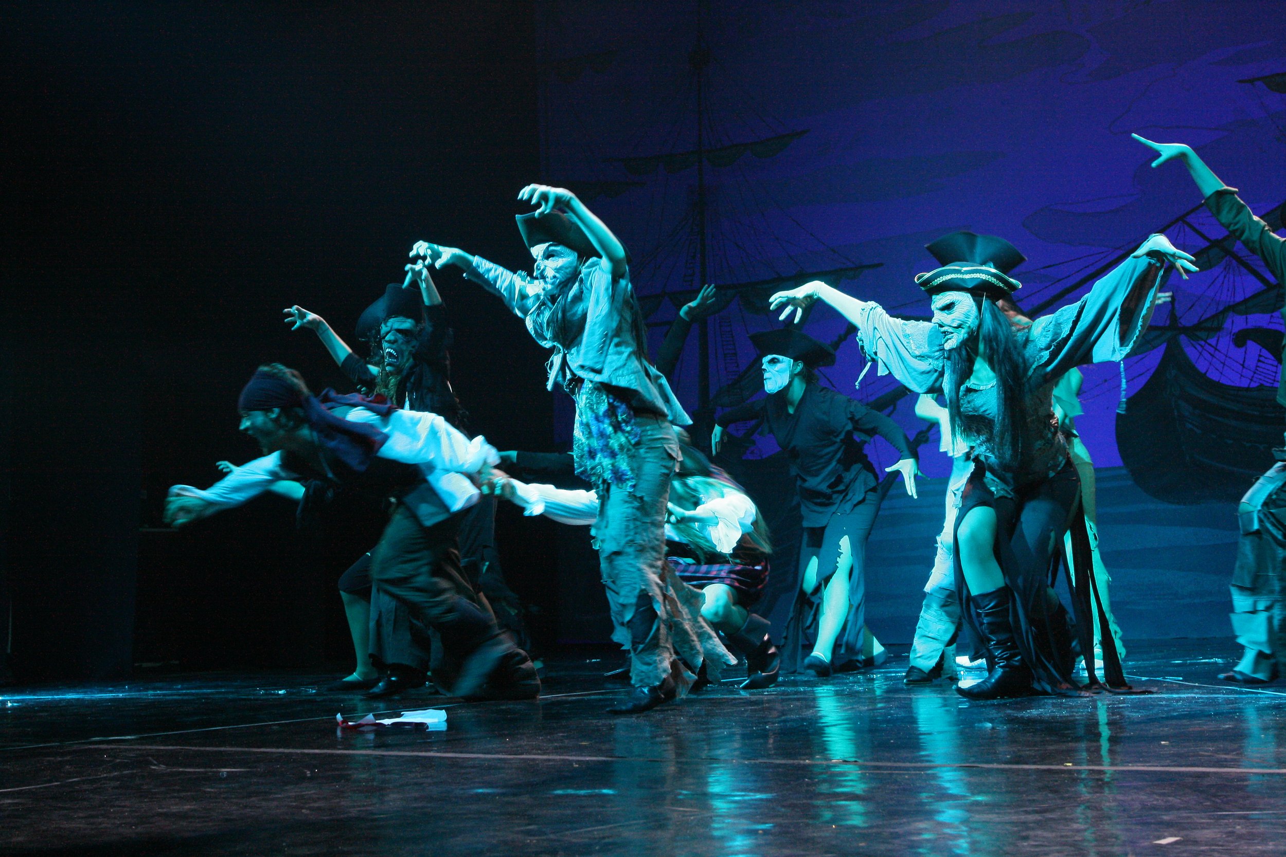 Performance teatrale con attori mascherati vestiti come pirates e pirati che danzano sul palco con uno sfondo di vele di una nave e luci blu.