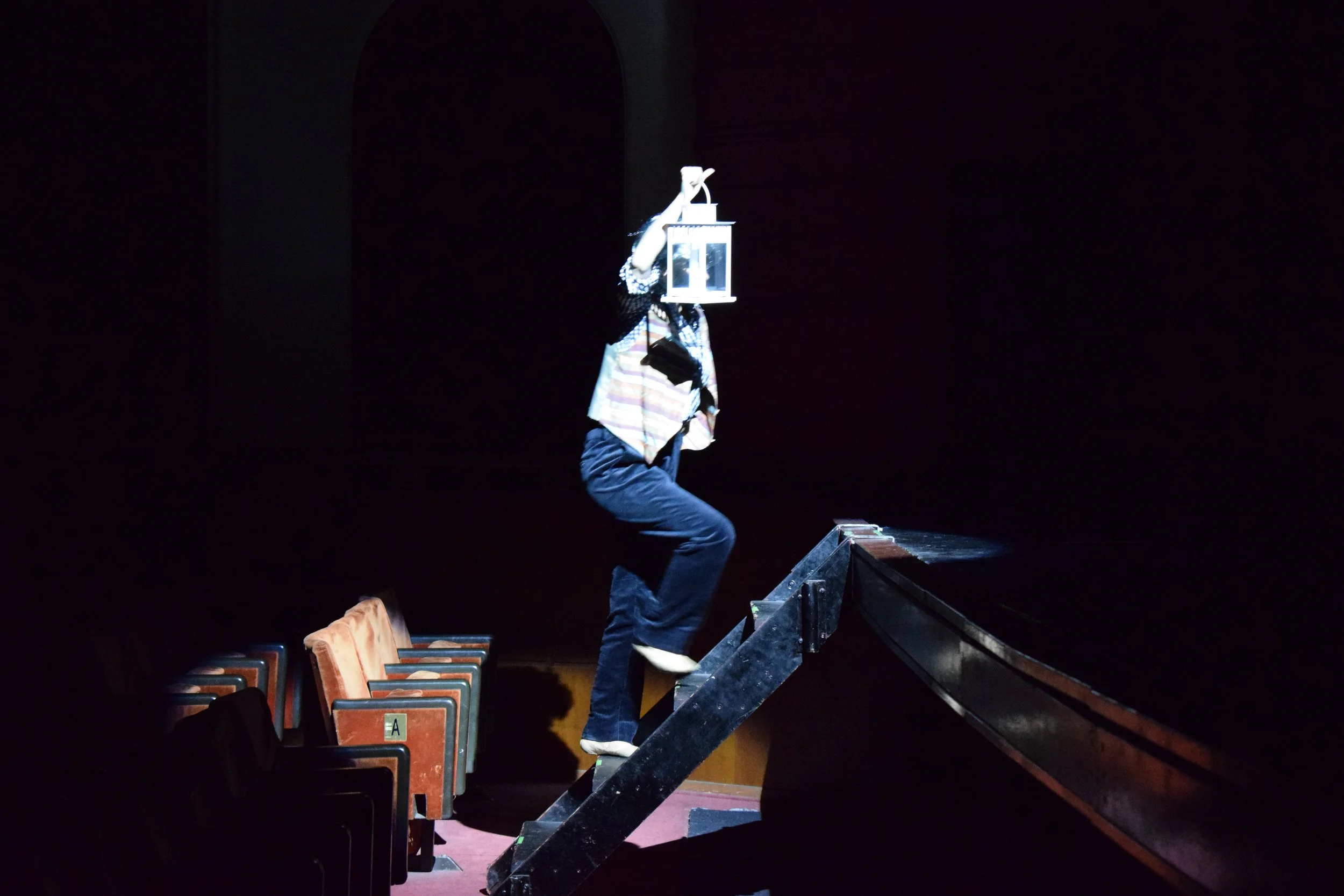Musicista in scena al teatro, salita sul palco, con strumenti musicali e sedie sullo sfondo, scena illuminata in modo drammatico.