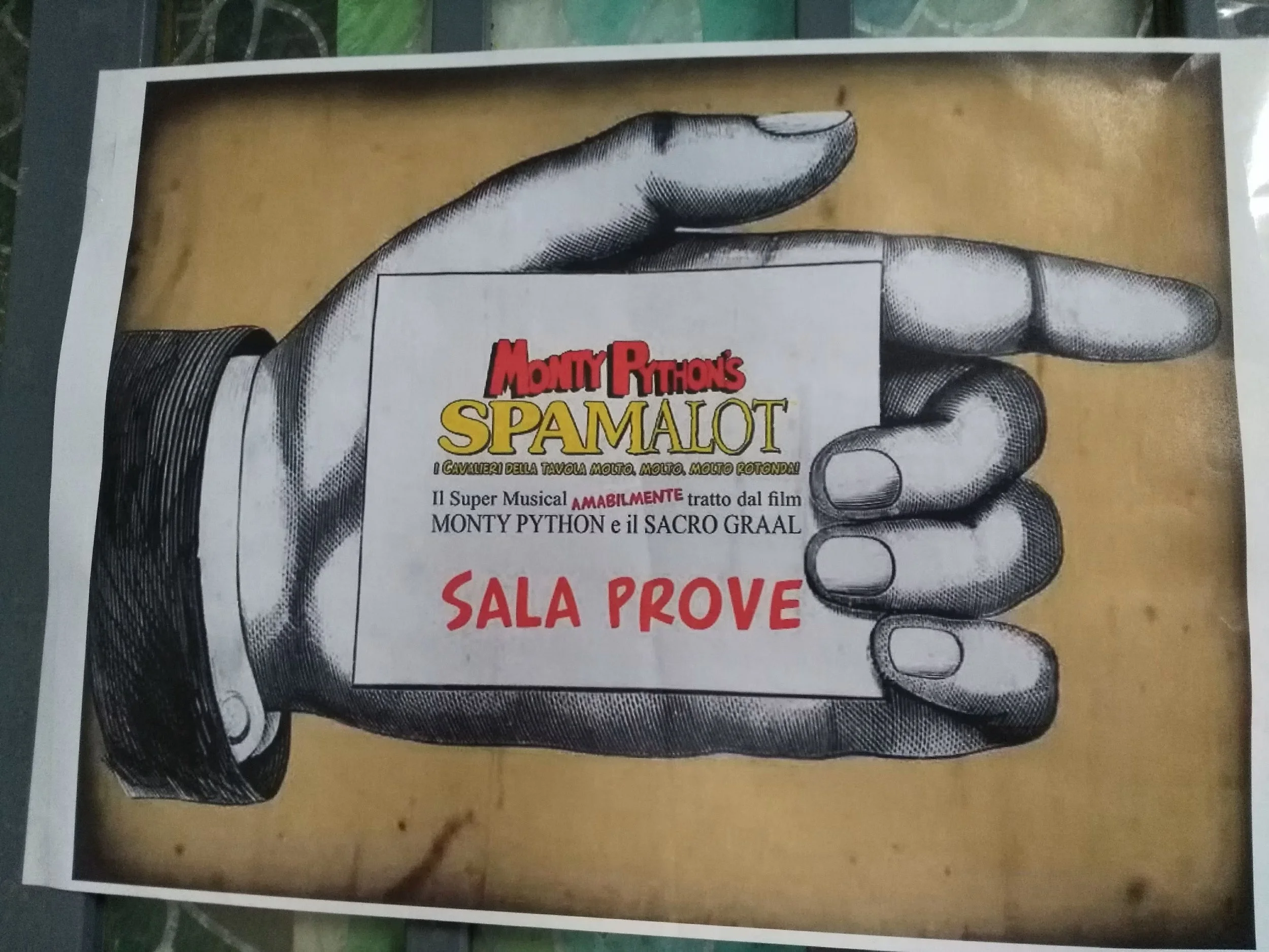 Poster con un disegno di una mano che tiene una scatola con scritto Monty Python Spamlot, promozione di un musical.