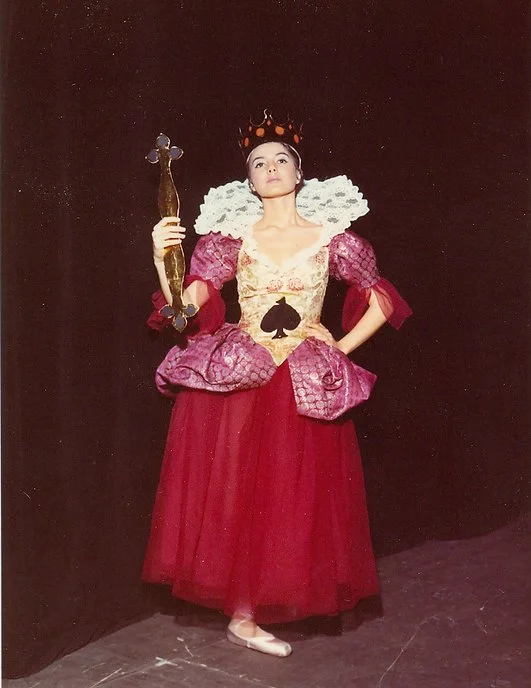 Donna in costume da regina, con una corona nera, abito rosso con dettagli dorati e paillettes, e una gilda di scacchi neri sulla gonna, che tiene in mano una quasi giocattolo. Sfondo nero.