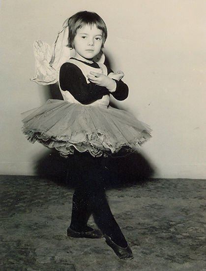 Un'immagine in bianco e nero di una bambina vestita come una ballerina con un tutù, calze nere e scarpette da ballo, mentre si mette in posa contro un muro vuoto.