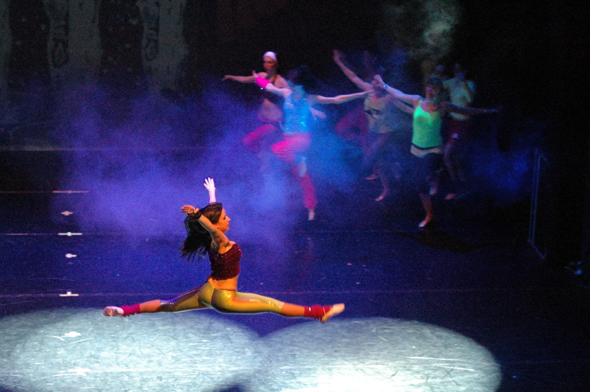 Un gruppo di ballerini in costume colorato eseguono una performance sul palco, alcuni in pose acrobatiche mentre altri sono in movimento.
