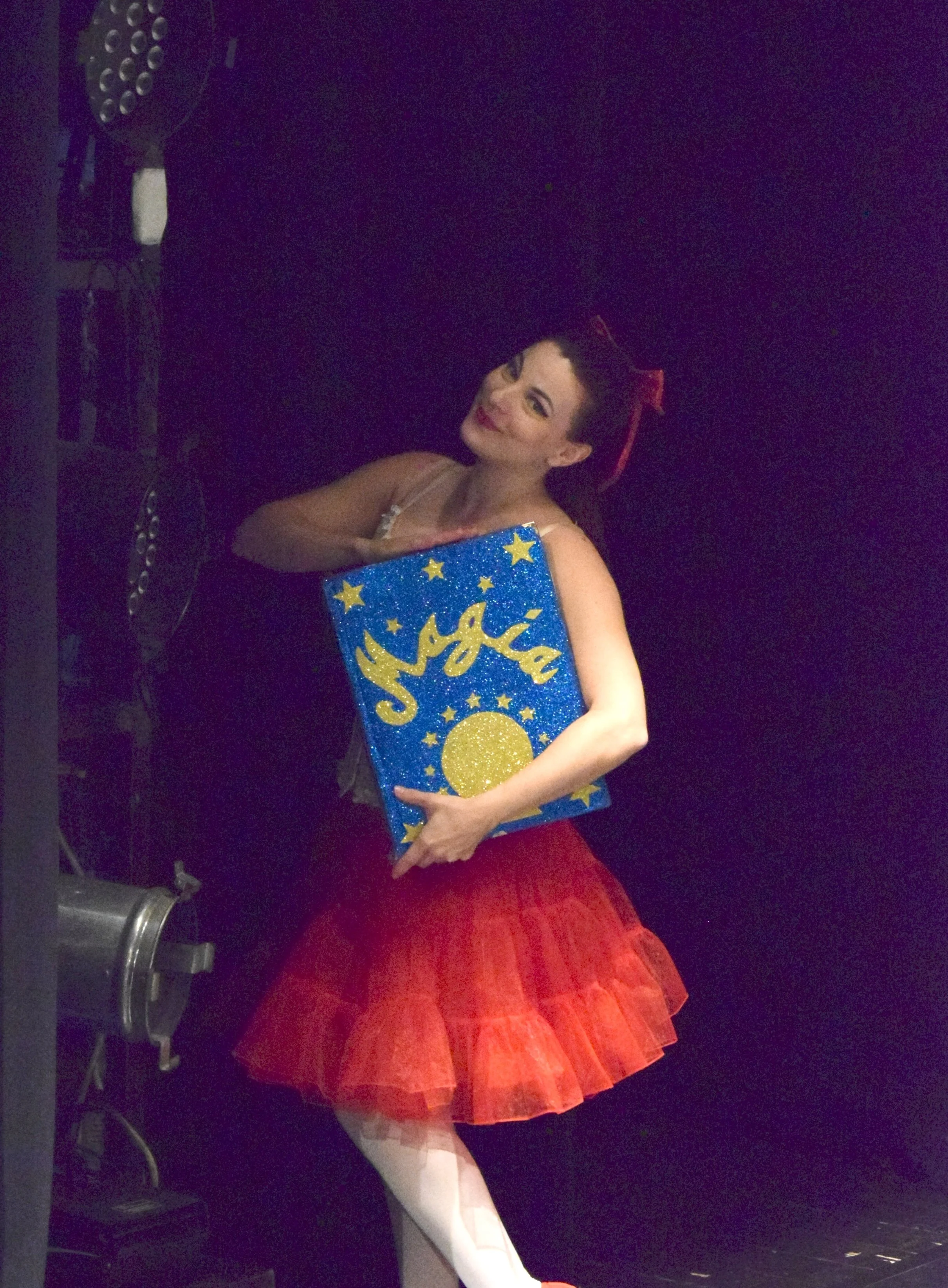 Una ballerina con una gonna rossa e un tutù bianco tiene un libro con una copertina blu decorata con stelle e un sole giallo. È in un teatro, con luci soffuse.