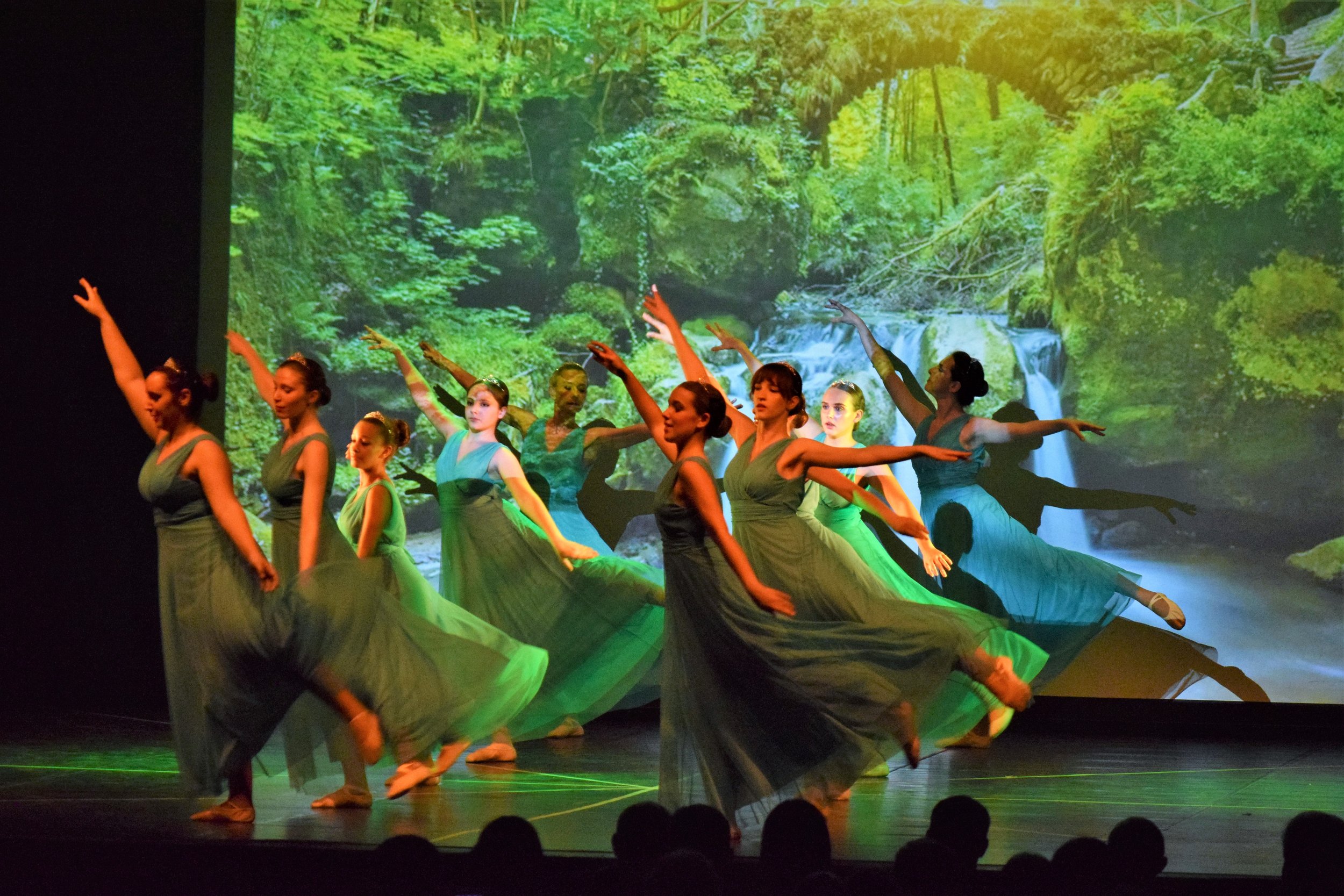 Gruppo di ballerine in costume verde che eseguono una coreografia su un palco con uno sfondo digitale di una foresta e una cascata.