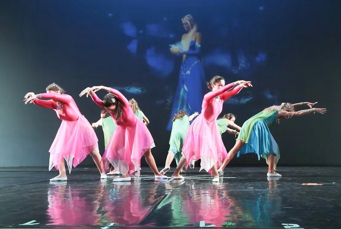 Gruppo di ballerine che danza sul palco, indossando costumi colorati, con uno sfondo con una proiezione di una donna in abito blu.