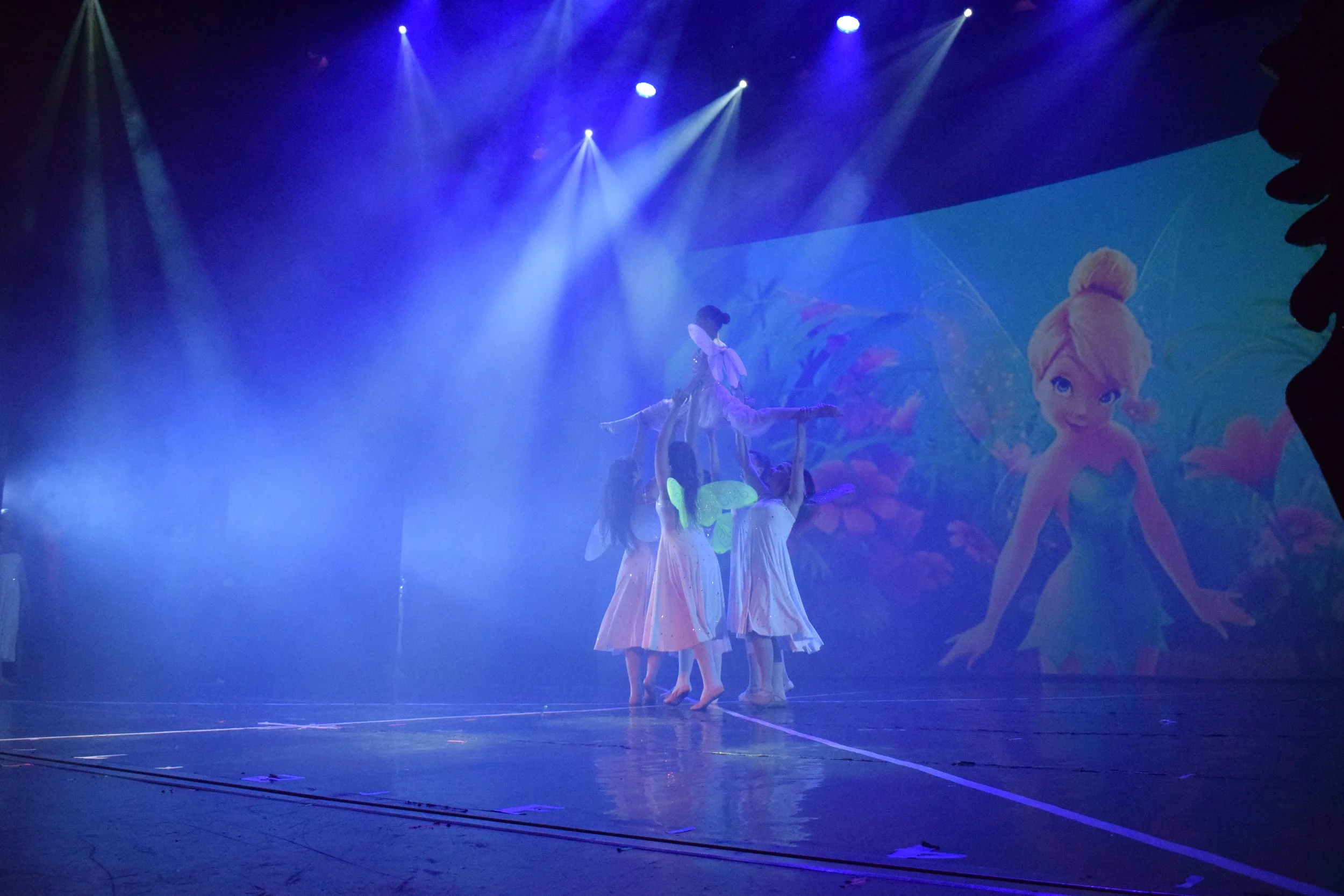Ragazze con costumi da fata che ballano sul palco durante uno spettacolo di danza, con uno sfondo luminoso e un grande schermo che mostra un personaggio di cartone animato.