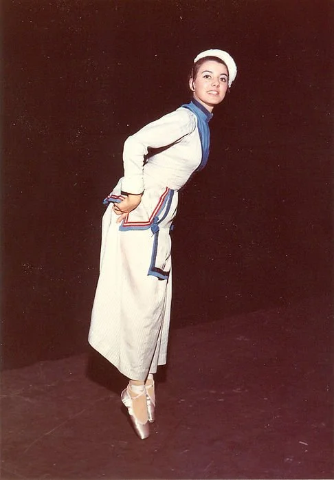Una ballerina posa in costume sul palco, indossa un tutù bianco con dettagli blu e una cuffia bianca, eseguendo una posizione di danza classica.
