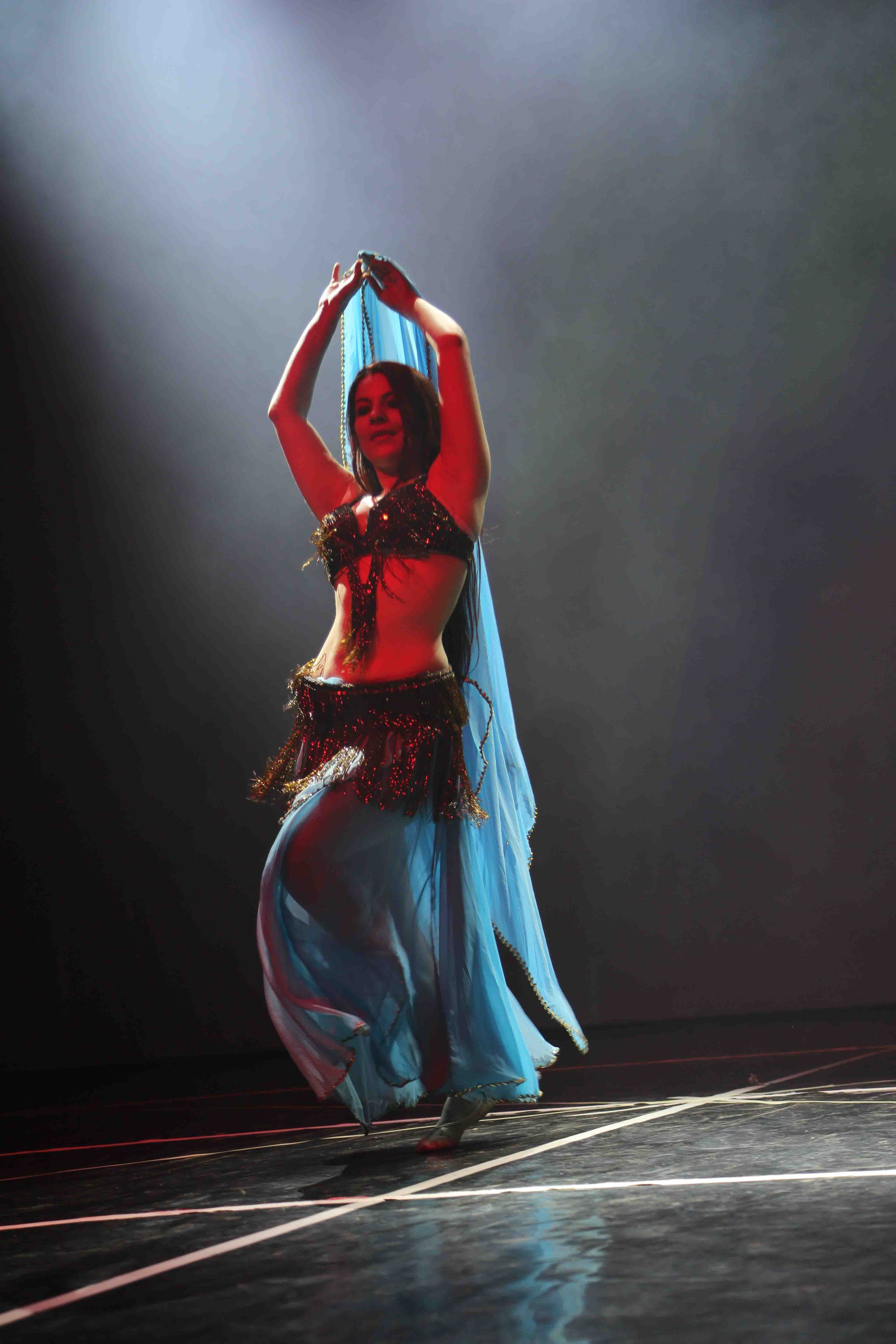 Una ballerina indossa un costume scintillante nero e oro, con una lunga gonna di tessuto blu, mentre danza sul palco sotto una luce soffusa.