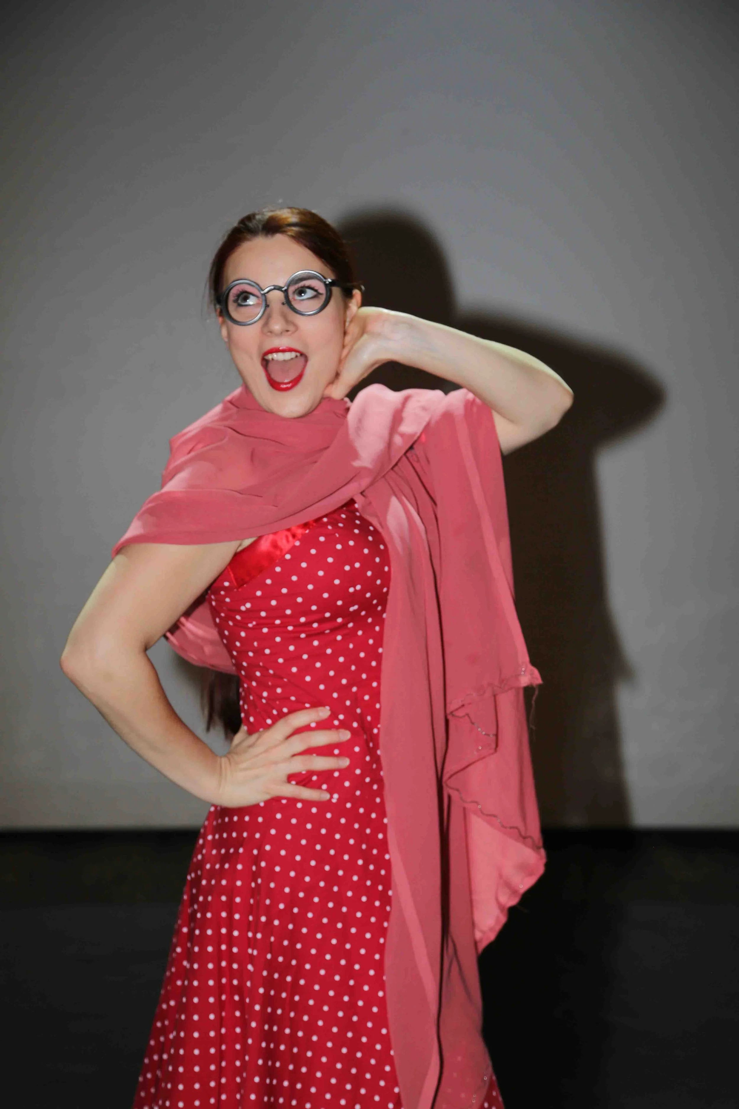 Una donna con occhiali spessi, capelli castani, vestita con un vestito rosso con pois bianchi e una sciarpa rosa, che sorride e posa con una mano sulla vita e l'altra dietro la testa, contro uno sfondo grigio.