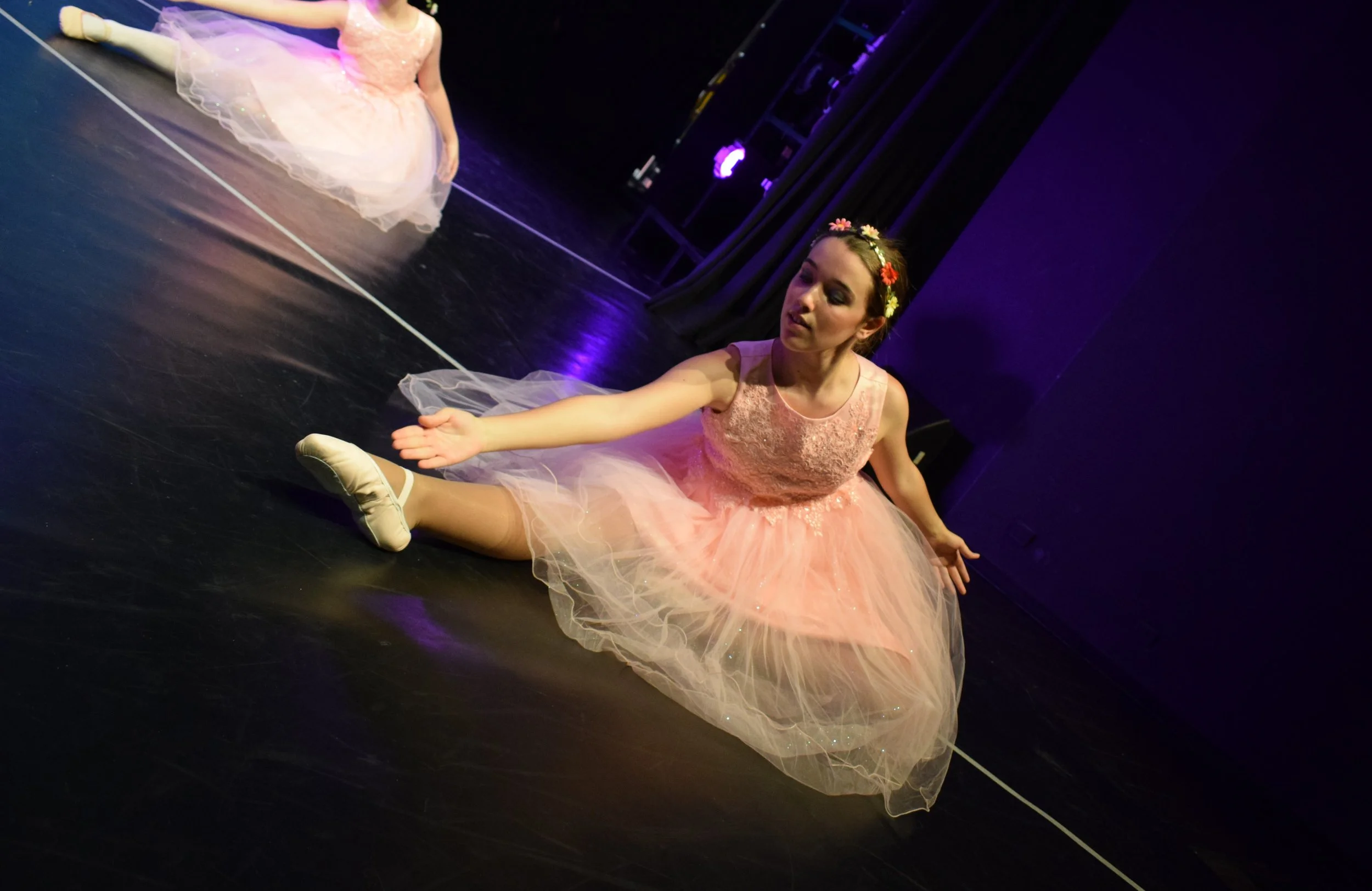 Bambina ballerina di balletto che si stira nel palco, indossa un delicato tutù rosa con fiorellini sulla testa, in un ambiente di teatro con luci soffuse.