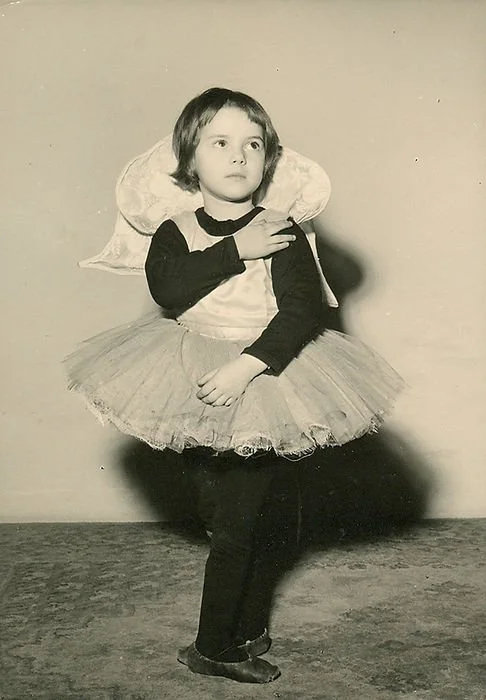 Una bambina in costume di ballerina con tutù e un grande fiocco sulla testa, in posa di fronte a uno sfondo semplice.