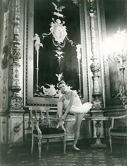 Una ballerina in costume bianco si sistema in una stanza elegante con arredi ricchi, pareti decorate con pannelli artistici e lampadari di cristallo.