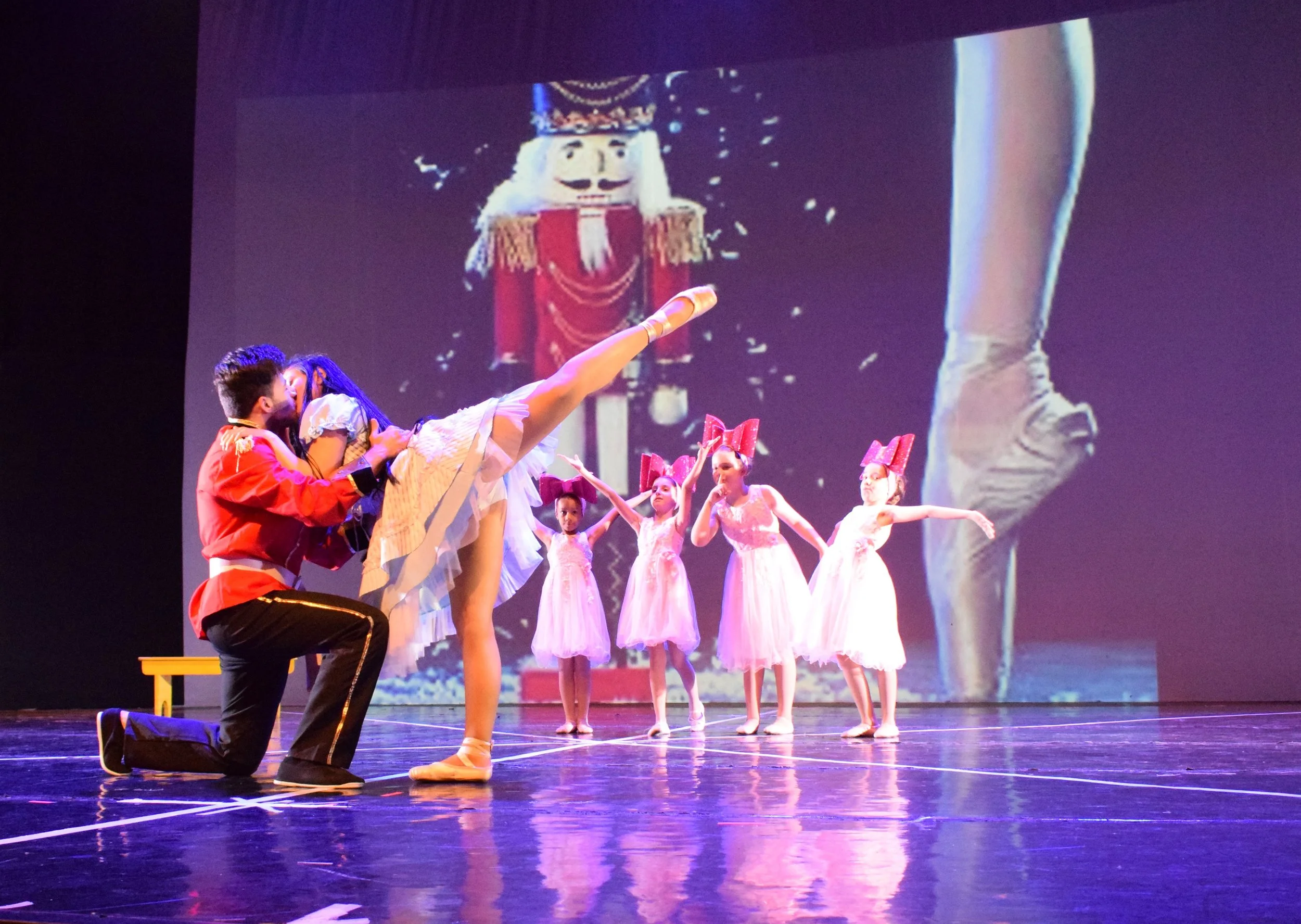 Una scena di un balletto con ballerine in costume rosa e persone in uniforme militare che si arrodano. Sullo sfondo, un grande schermo mostra un soldato con barba e cappello storico.