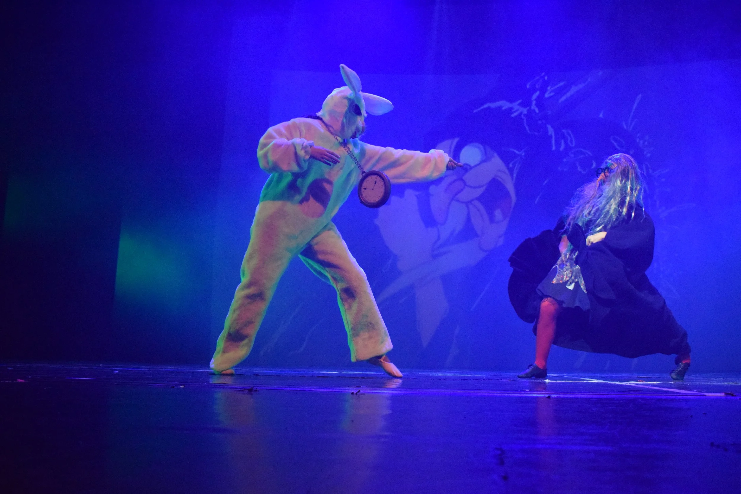 Una scena di teatro con due persone in costume. Una persona con costume da coniglio con orologio appeso al collo e una persona con parrucca lunga e vestiti eleganti, sullo sfondo un disegno di un coniglio.