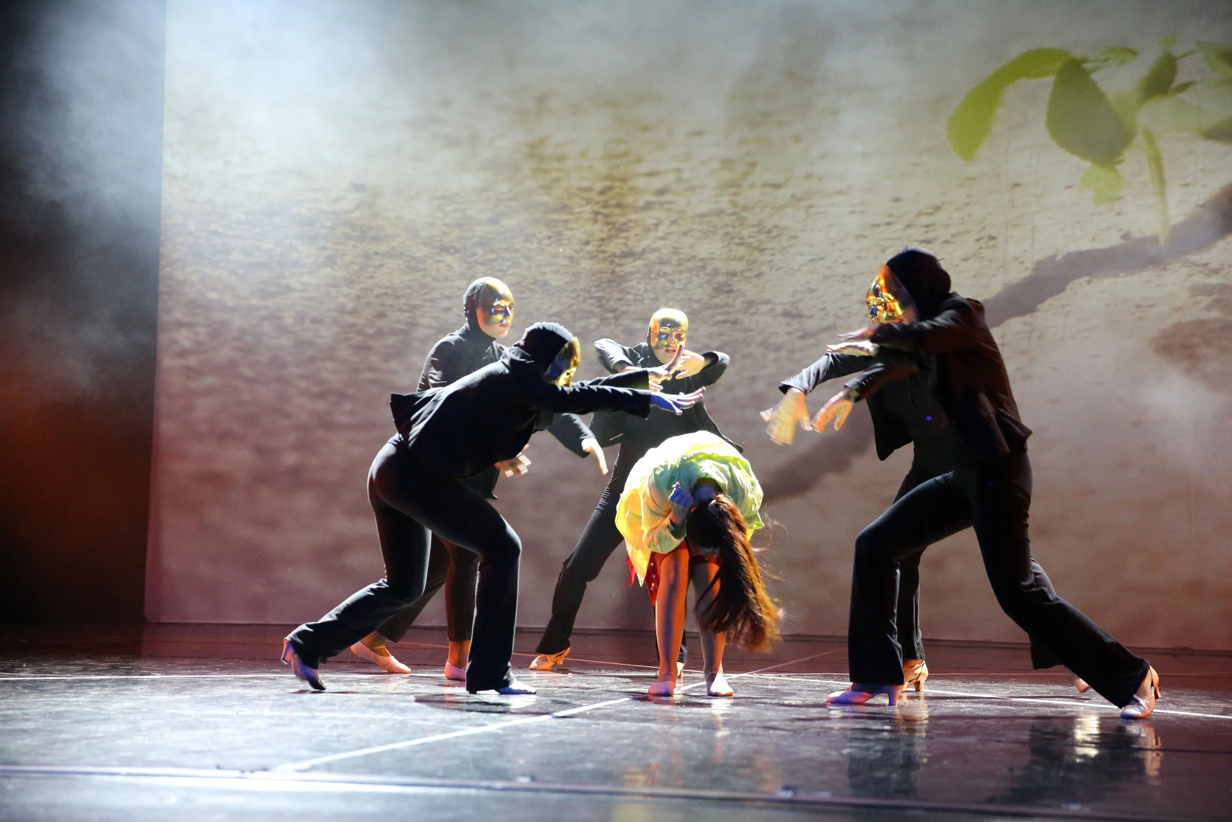 Un gruppo di ballerini indossa costumi neri con maschere dorate e si esibiscono in una coreografia su un palco illuminato con uno sfondo che mostra un'immagine di foglie verdi.