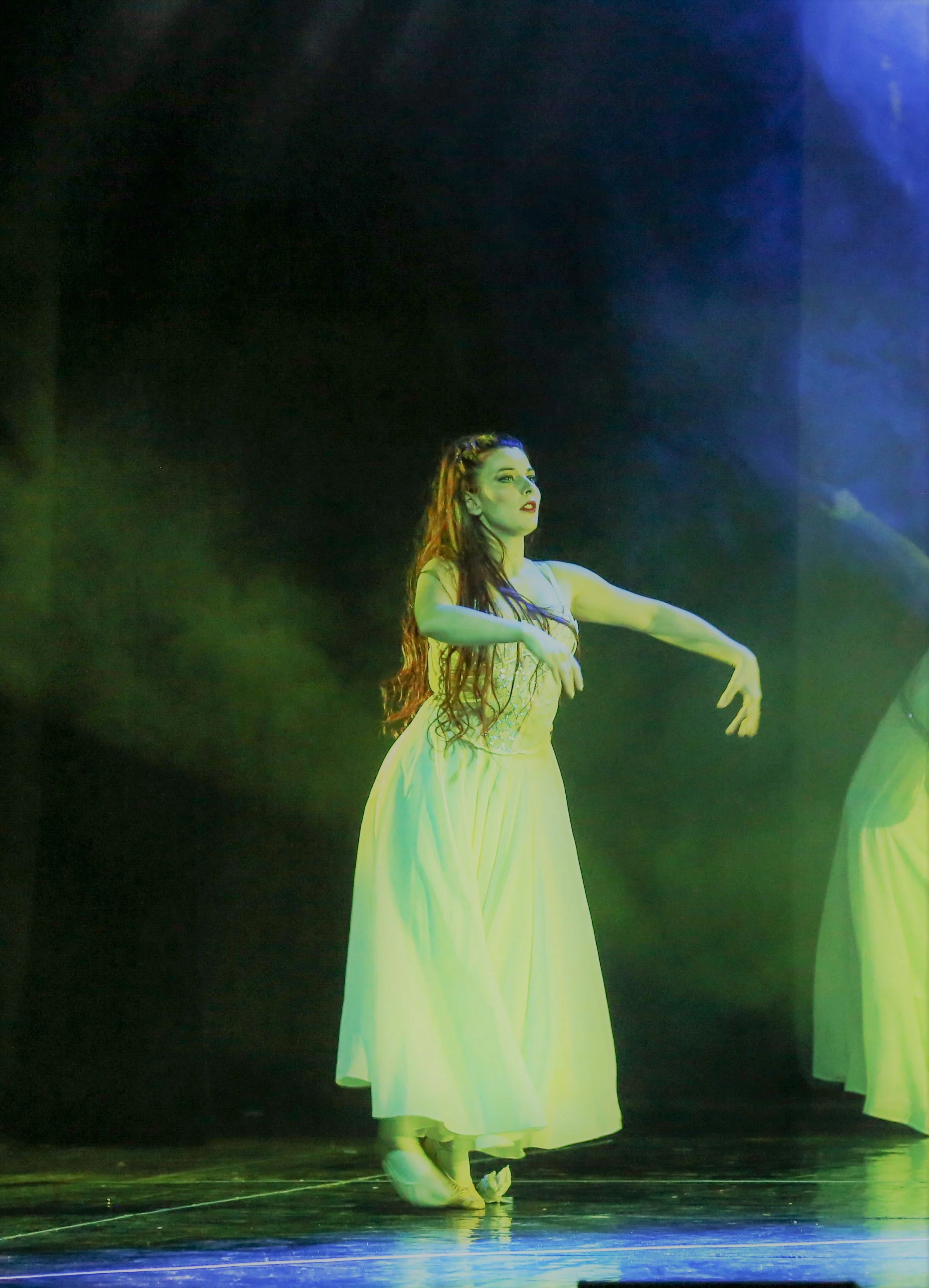 Ballerina con vestito lungo bianco, danza sotto luci verdi e blu in un teatro.