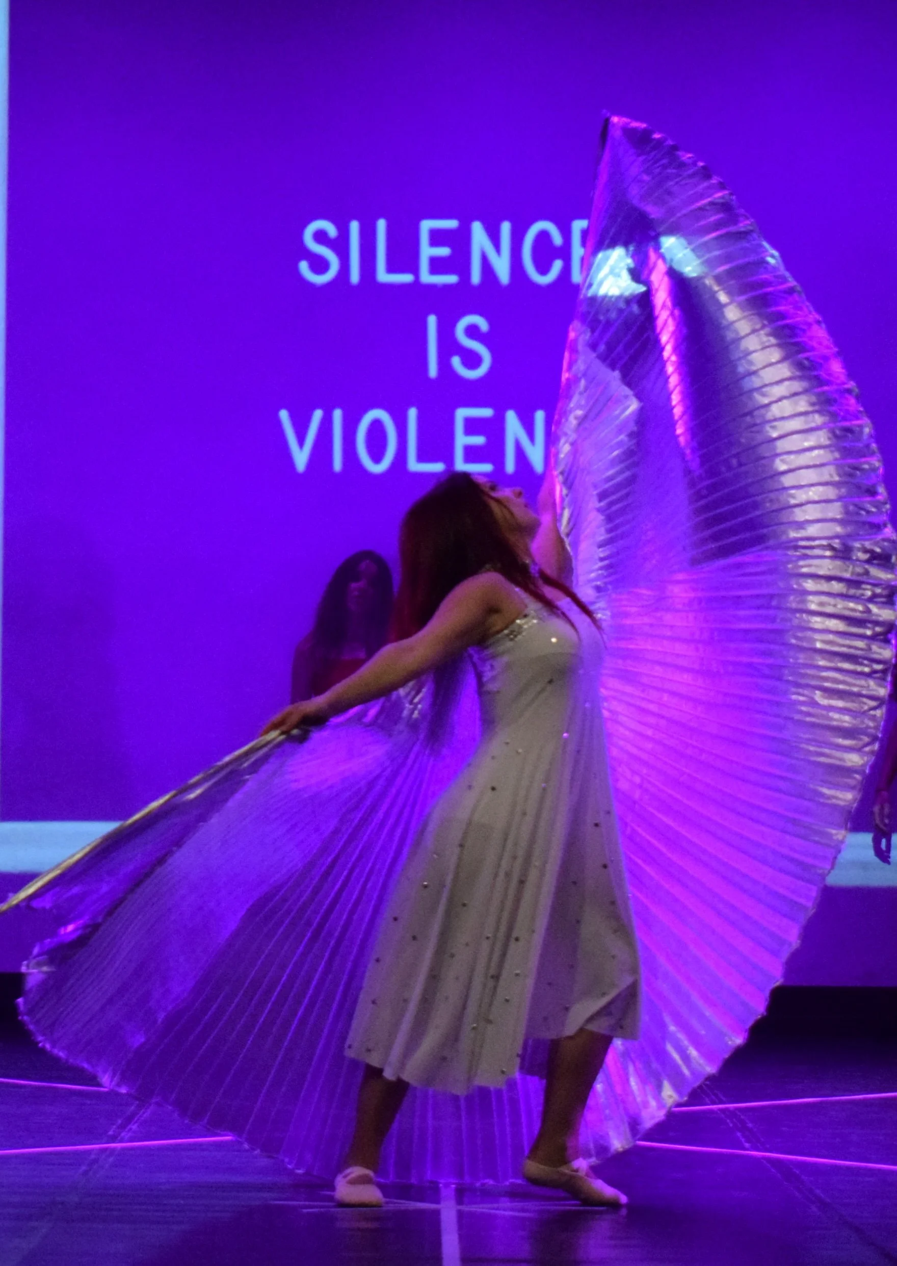 Una ballerina con un abito lungo, bianco con dettagli dorati, e un mantello pieghettato gigante oro semi-trasparente illuminato di viola, si esibisce sul palco. Sullo sfondo si vede la scritta 'Silence is Violent' in luce blu.