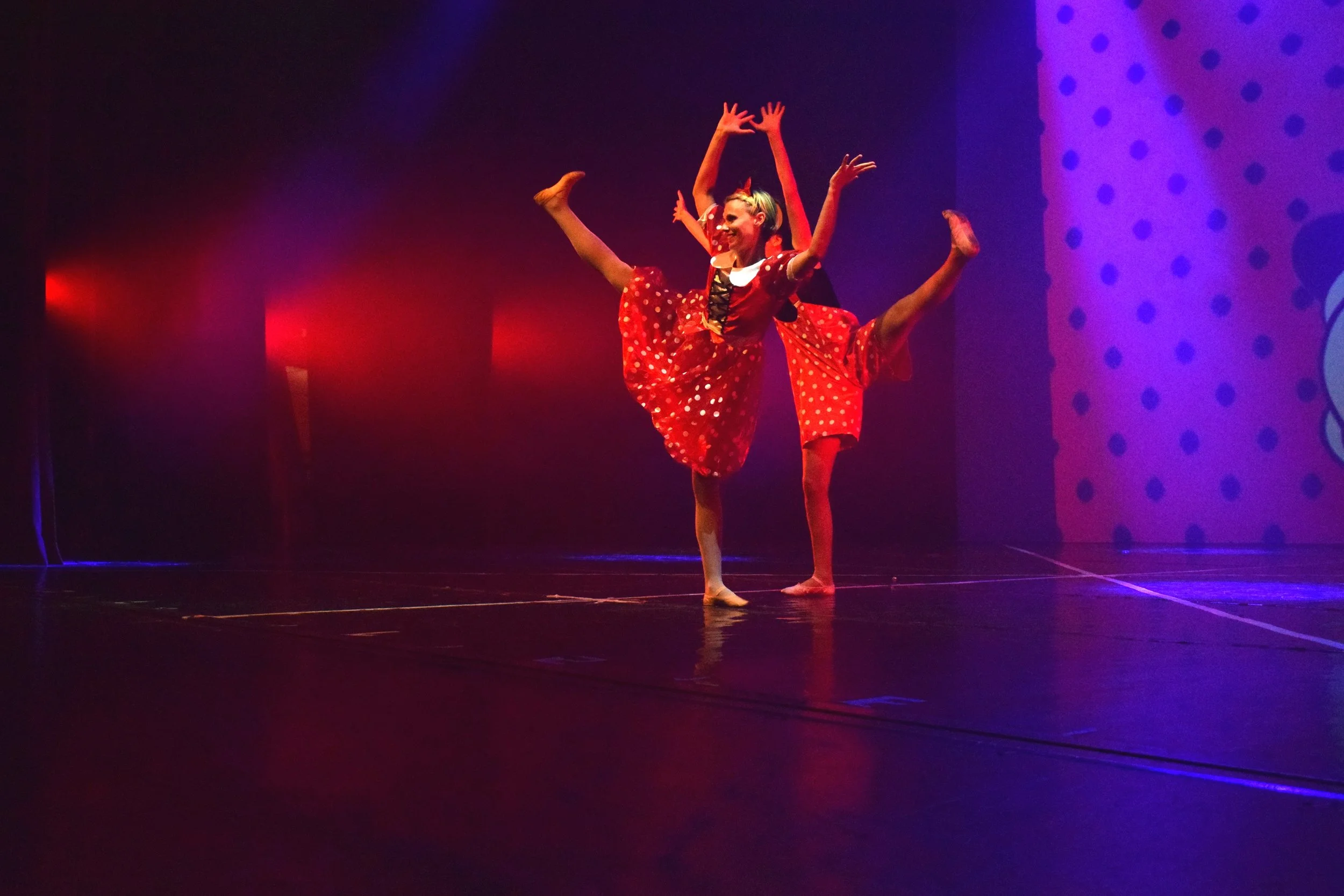 Due ballerine vestite con abiti rossi a pois bianchi mentre eseguono una coreografia sul palco con luci colorate