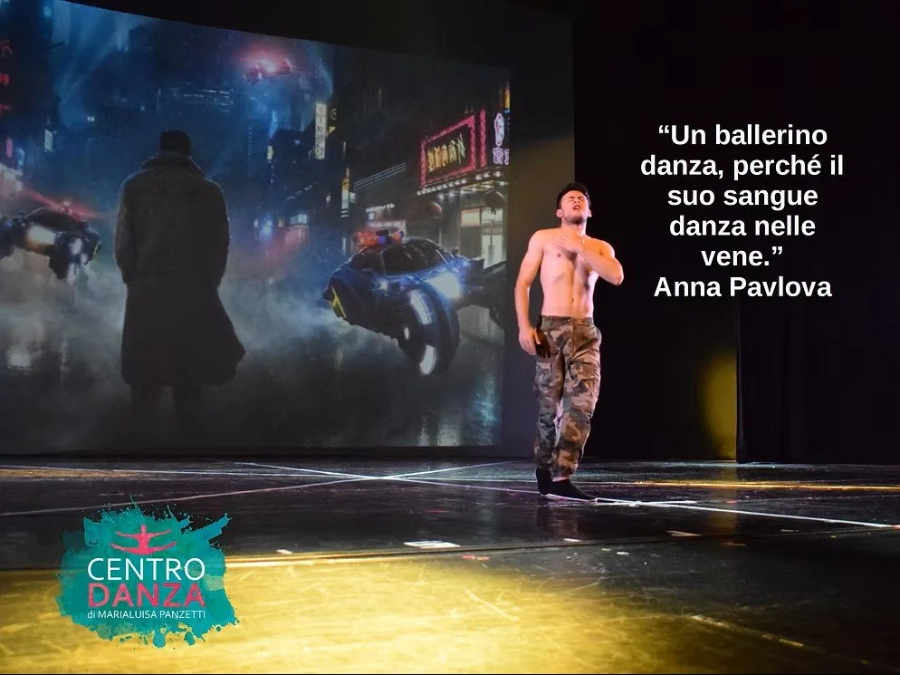 In scena, un ballerino maschio senza maglia, con pantaloni cargo militari, posizionato a destra di fronte a uno sfondo di città futuristica con luci neon e veicoli volanti. Sullo sfondo, una citazione di Anna Pavlova: "Un ballerino danza, perché il suo sangue danza nelle vene." Sul lato sinistro, il logo di un centro danza.