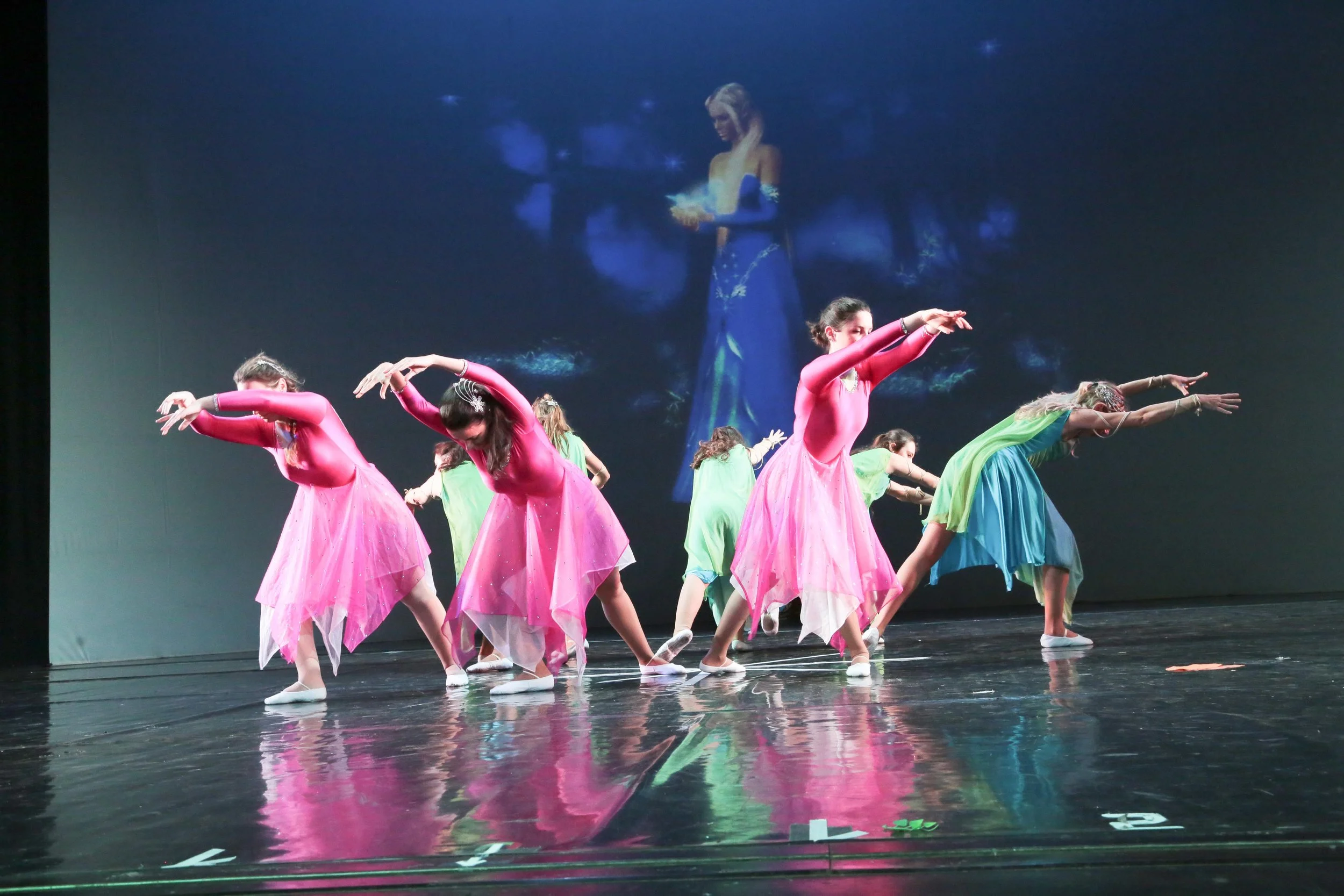 Un gruppo di ballerine indossa costumi colorati rosa, verde e blu mentre eseguono una coreografia sul palco, con una proiezione di una donna in abito blu sullo sfondo.