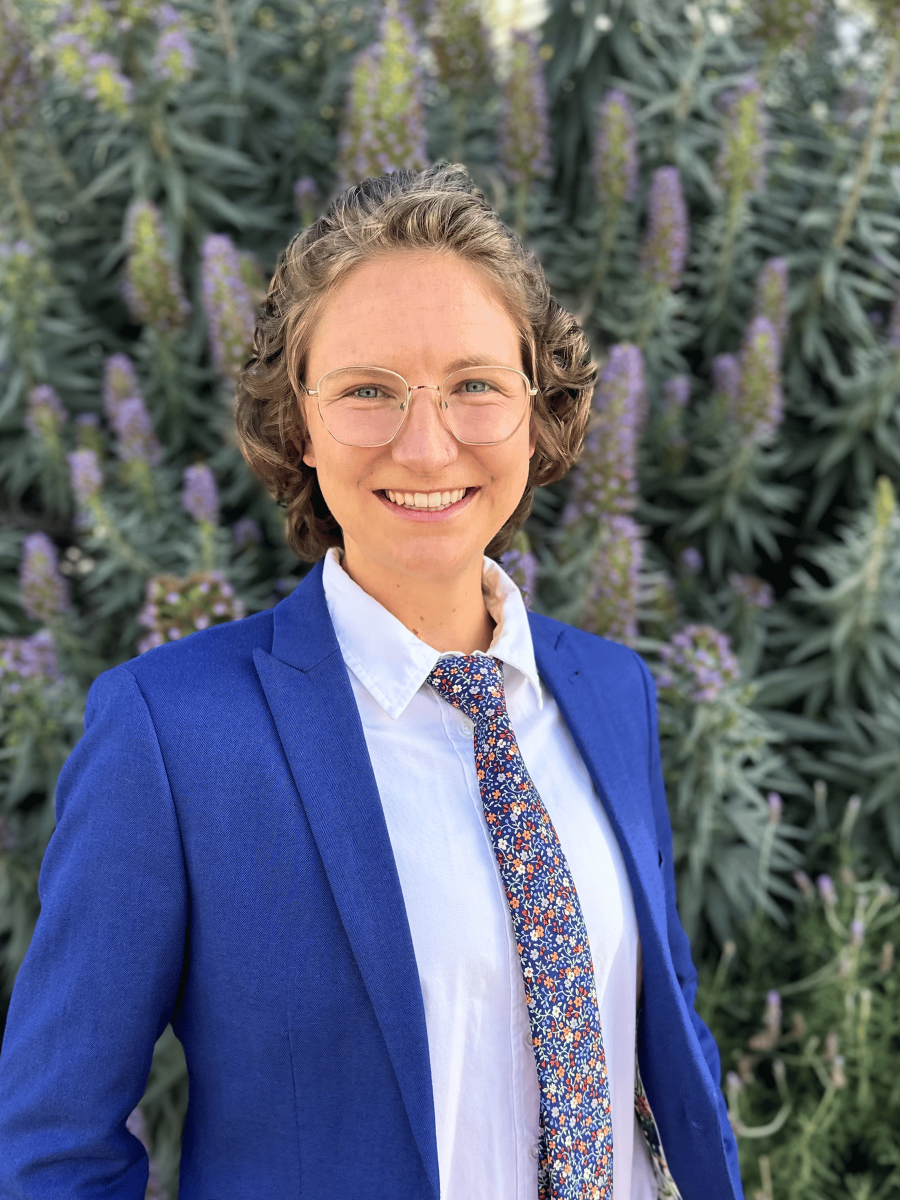 A woman with short curly hair, glasses, and light skin, smiling in front of a background of purple flowering plants. She is wearing a blue blazer, a white shirt, and a colorful patterned tie.