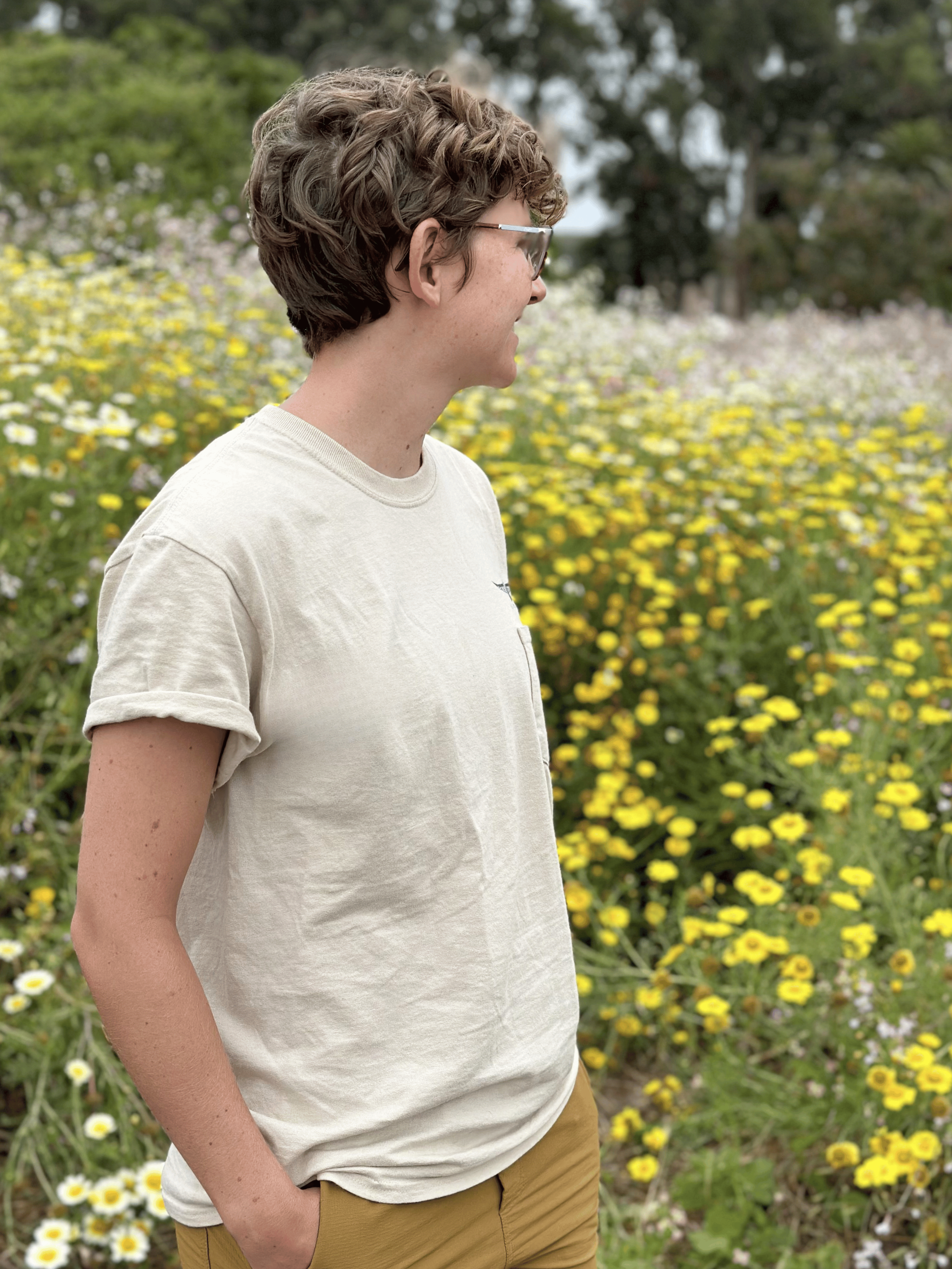 Side view of a person with curly short hair and glasses, wearing a light-colored T-shirt and khaki pants, standing outdoors in a field of yellow and white flowers with trees in the background.