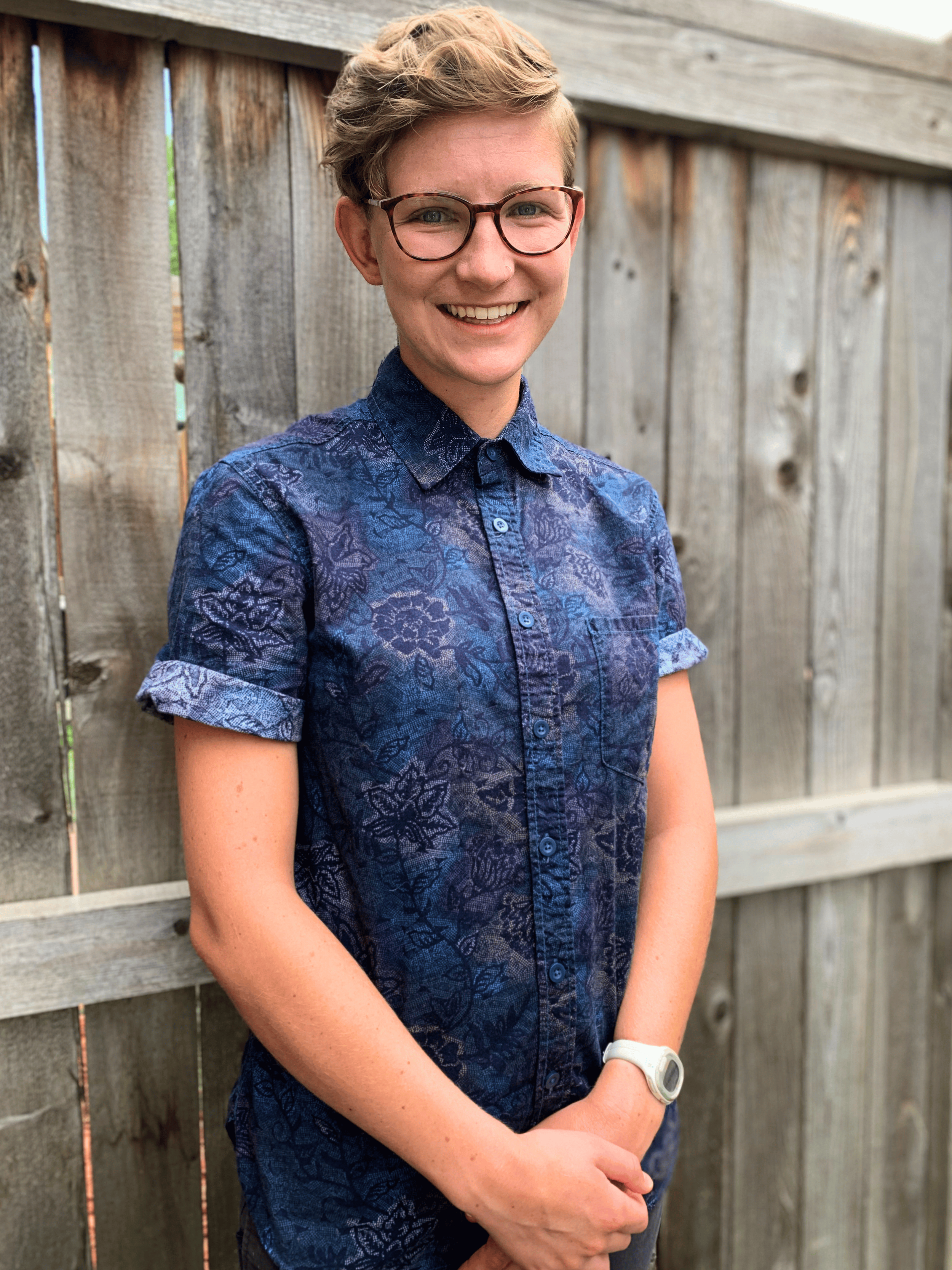 A smiling woman with short curly blond hair and glasses standing in front of a wooden fence, wearing a blue patterned button-up shirt and a white watch.