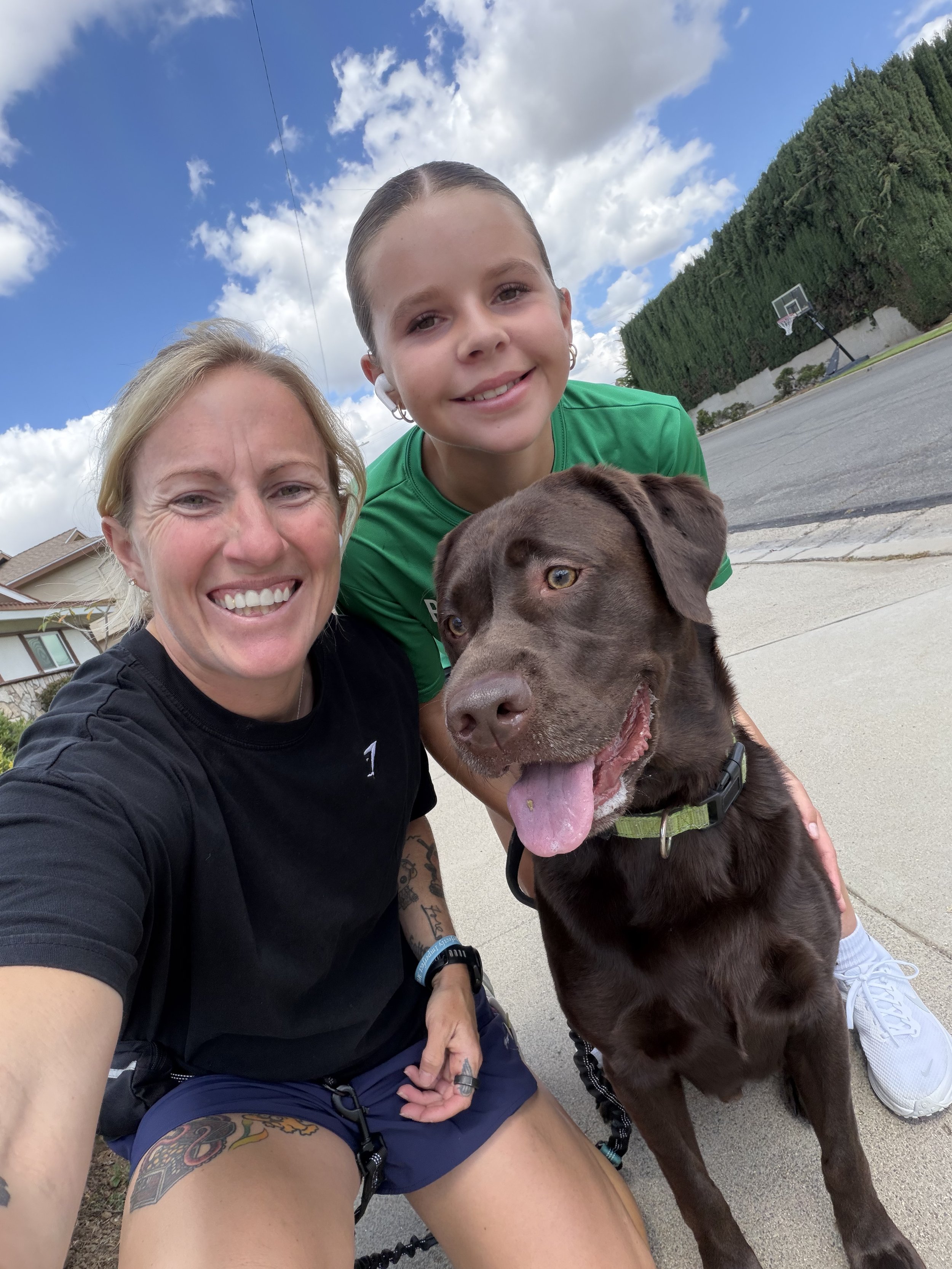 A woman, a young girl, and a large brown dog outdoors on a sidewalk, with a basketball hoop and trees in the background. The woman is smiling at the camera, wearing a black shirt and blue shorts, and has tattoos and bracelets on her arm. The girl is also smiling, wearing a green shirt and white shoes, and is holding the dog. The dog is sitting, has its tongue out, and is looking slightly to the side.