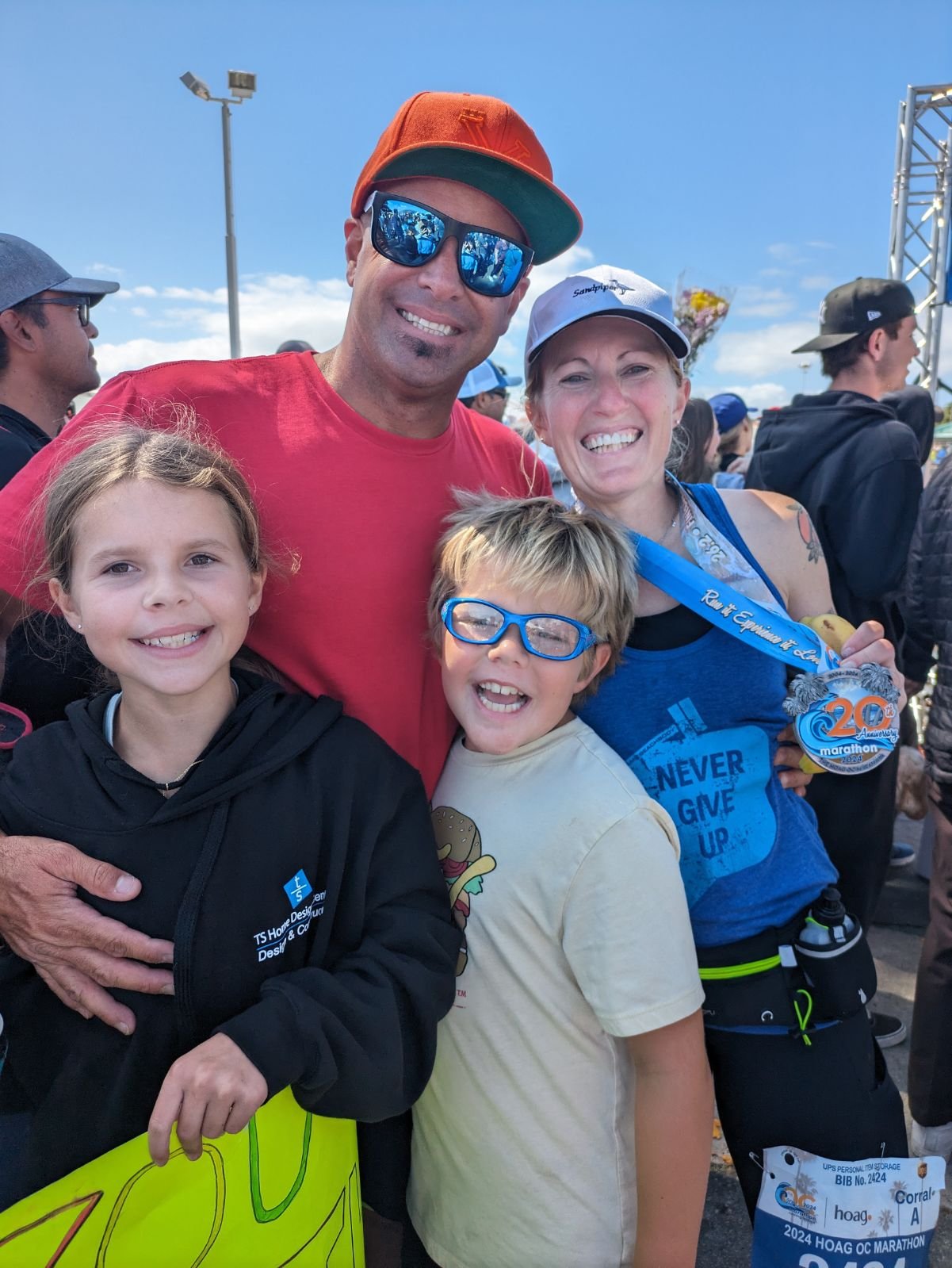 A family celebrating after completing a marathon, with a man and a woman holding medals and children smiling in front of a crowd outdoors.