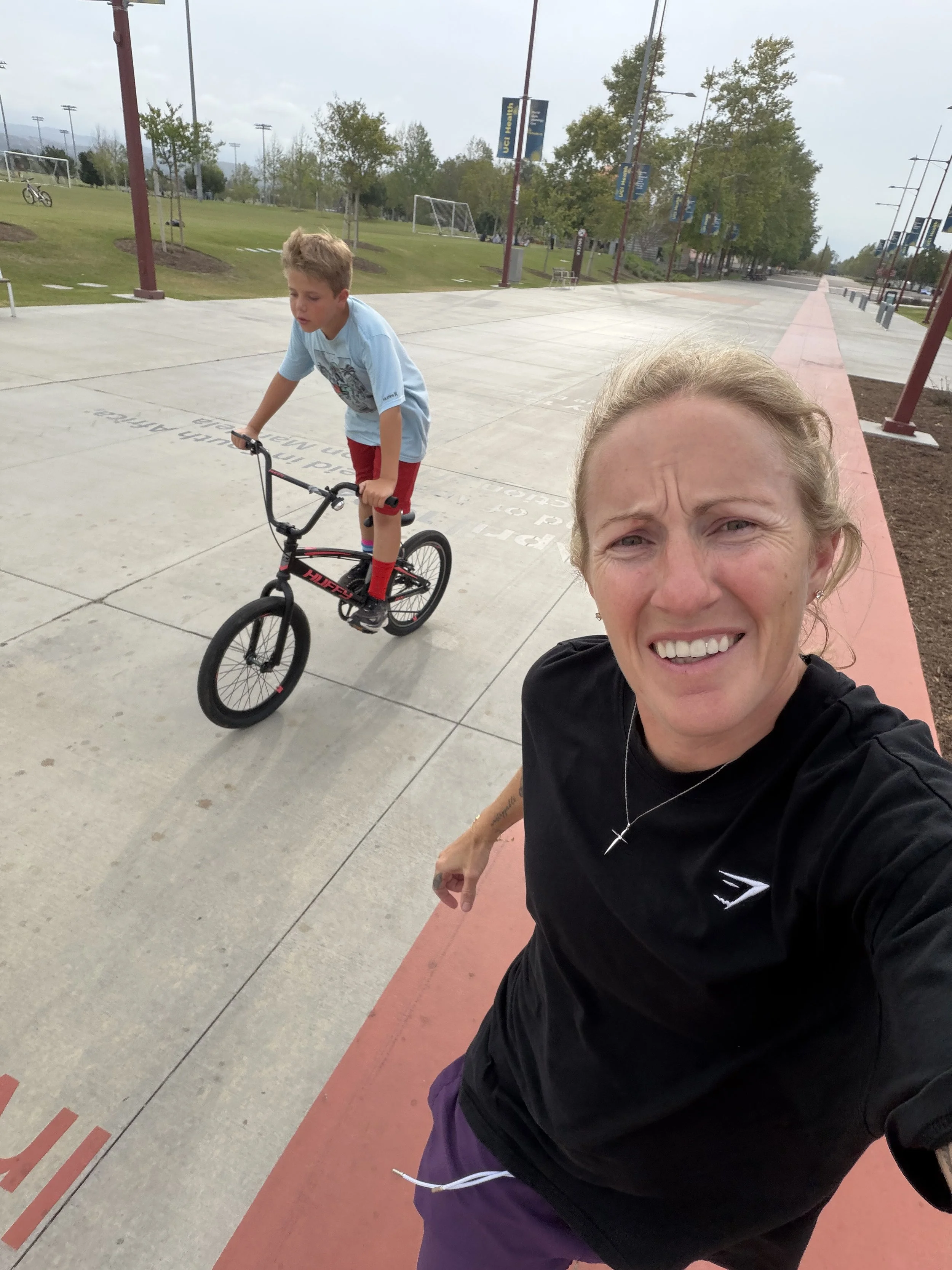 A woman taking a selfie outdoors at a park, smiling and showing her teeth, with a young boy riding a BMX bike in the background on a concrete pathway.