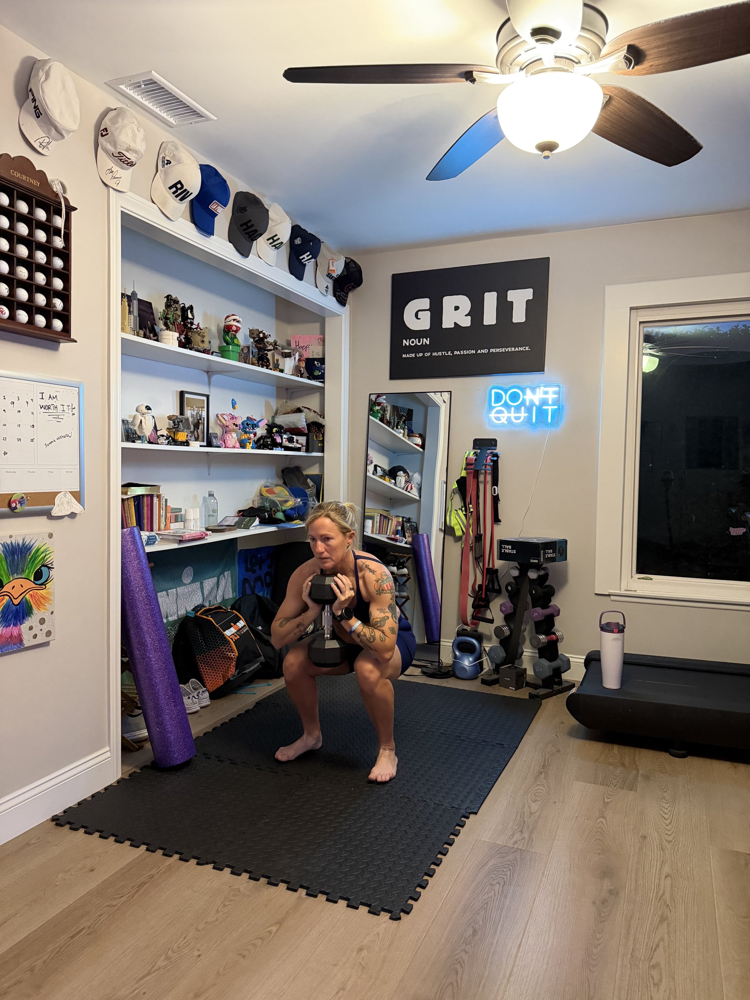 A woman performing a squat exercise while holding a dumbbell in a home gym. The room has a ceiling fan, a black workout mat, a treadmill, shelves with toys and caps, and motivational signs on the wall.
