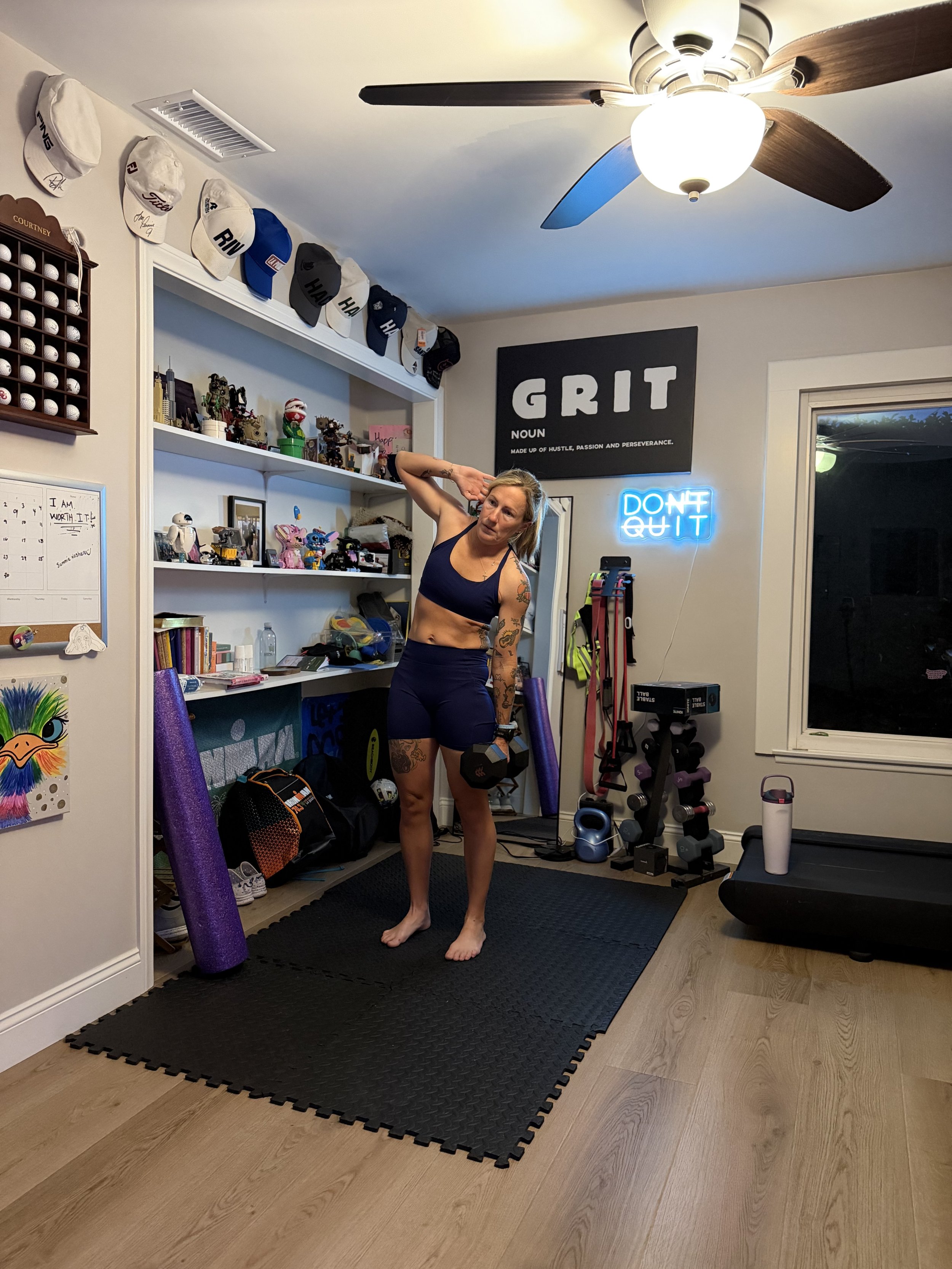 A woman standing in a home gym, holding a dumbbell, surrounded by workout equipment, and decorated with motivational signs and memorabilia.