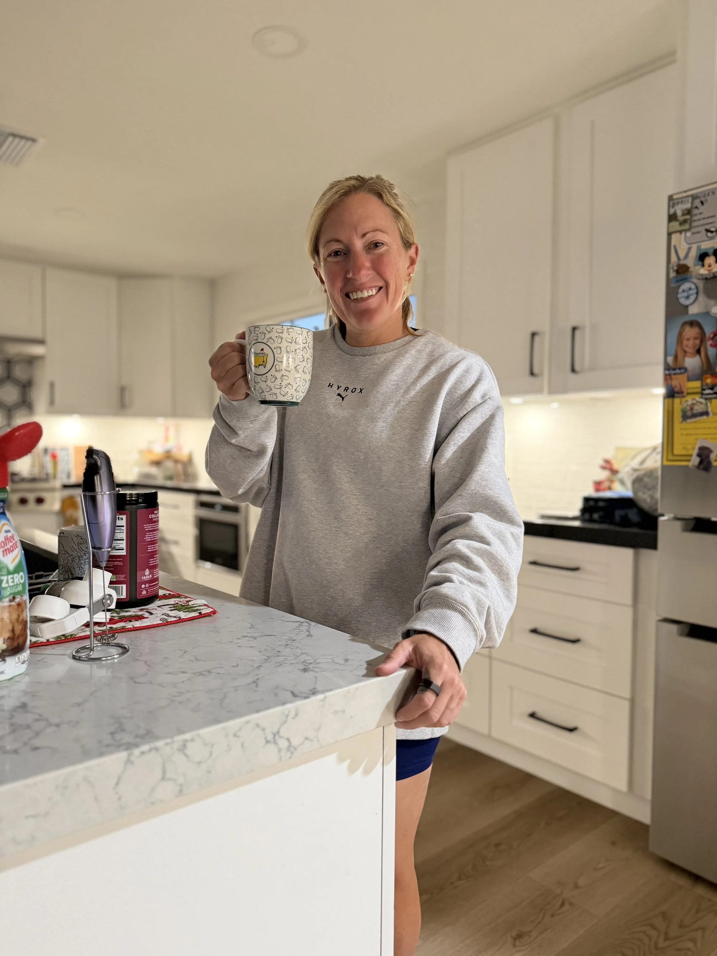 A smiling woman with blonde hair, wearing a gray sweatshirt and shorts, holding a mug in her right hand, standing in a modern kitchen.