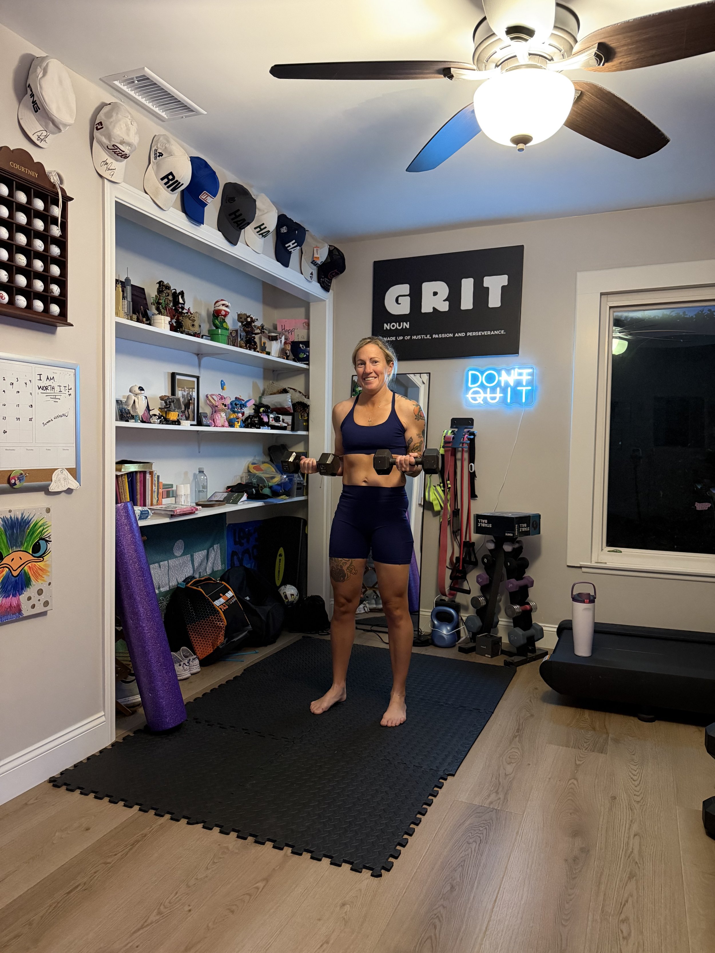 A woman in workout clothing standing in a home gym holding dumbbells, with exercise equipment, motivational signs, and hats on a shelf behind her.