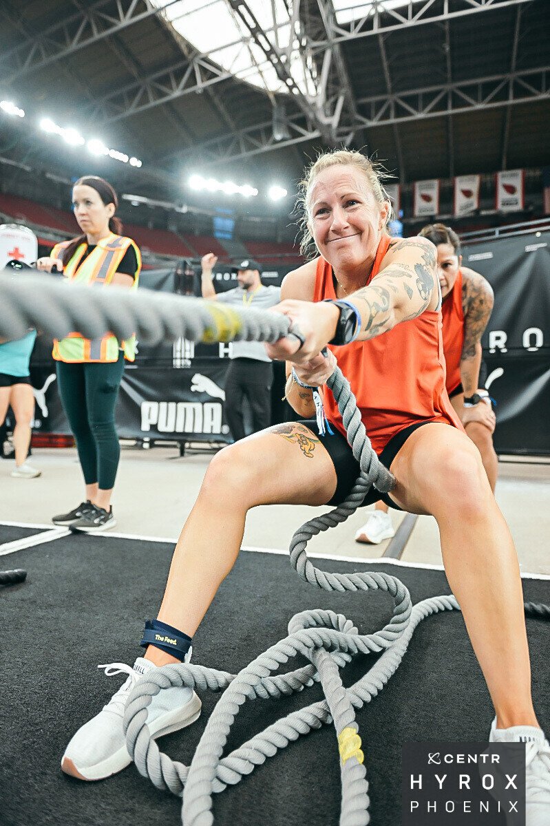A woman with tattoos on her arms and leg is participating in a battle rope workout at an indoor sports arena. She is wearing an orange sleeveless top, black shorts, and white athletic shoes, and appears to be exerting effort. Other individuals and a large banner with the Puma logo are visible in the background.