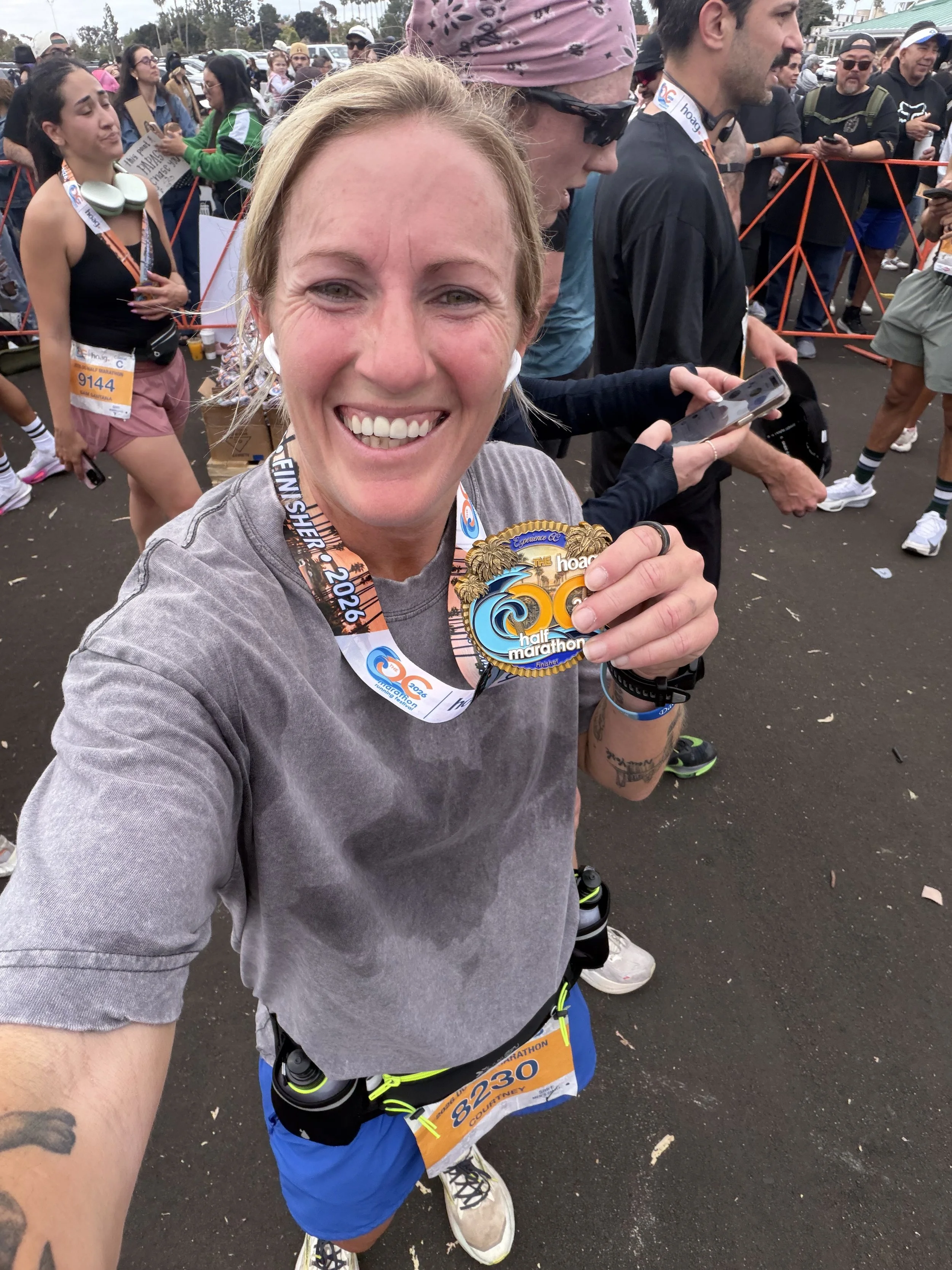 A woman smiling for a selfie after finishing a half marathon, holding her medal. She is surrounded by other runners and spectators at the event.