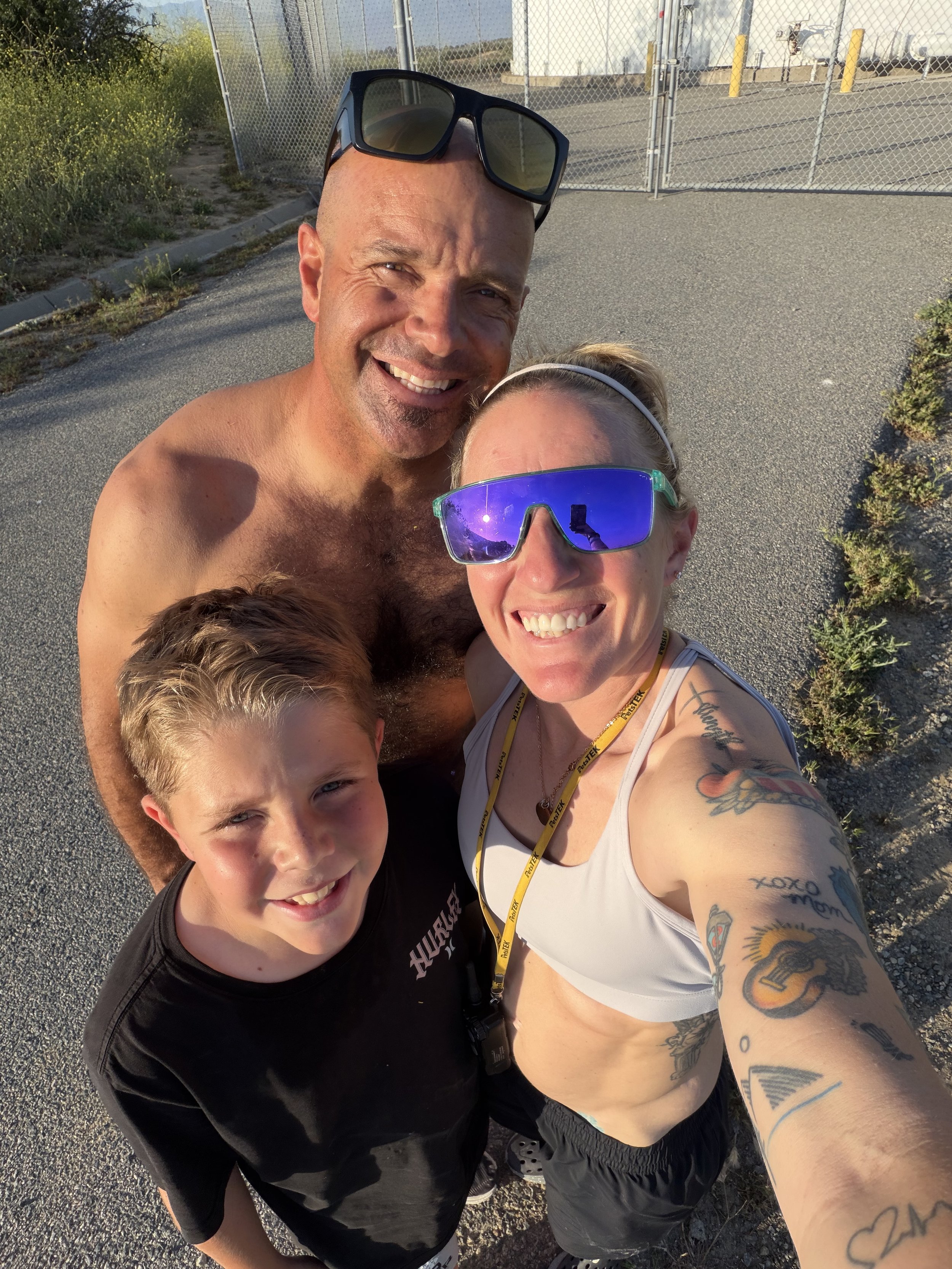 A group selfie of four smiling people outdoors near a chain link fence and parking lot, taken in bright sunlight. Two are adults, one man and one woman, with the woman wearing sunglasses and visible tattoos on her arm, and two children, one with blonde hair and one with brown hair.