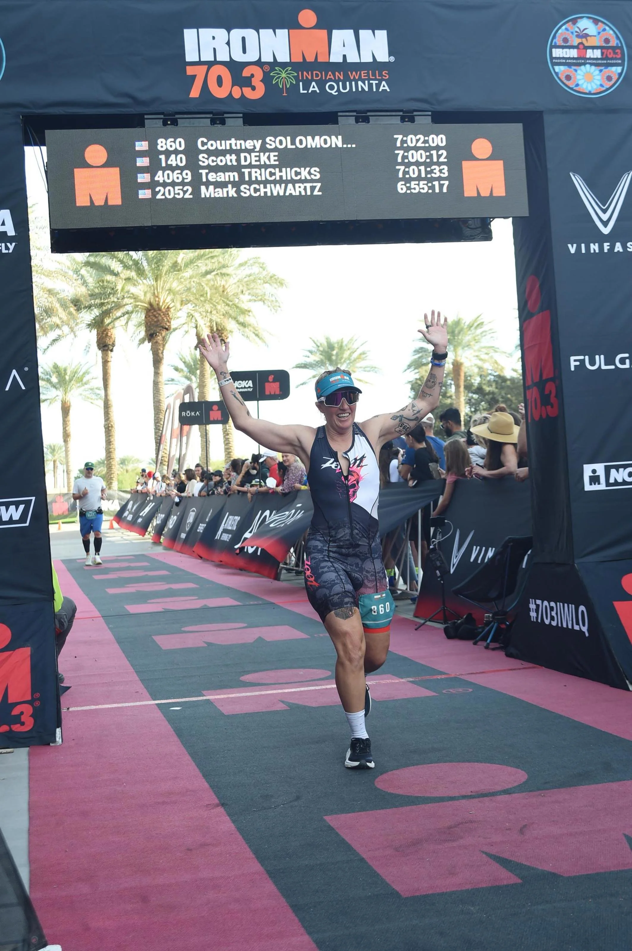 Female athlete crossing the finish line at an Ironman race, smiling and raising her arms in victory, with a digital timer and spectators in the background.