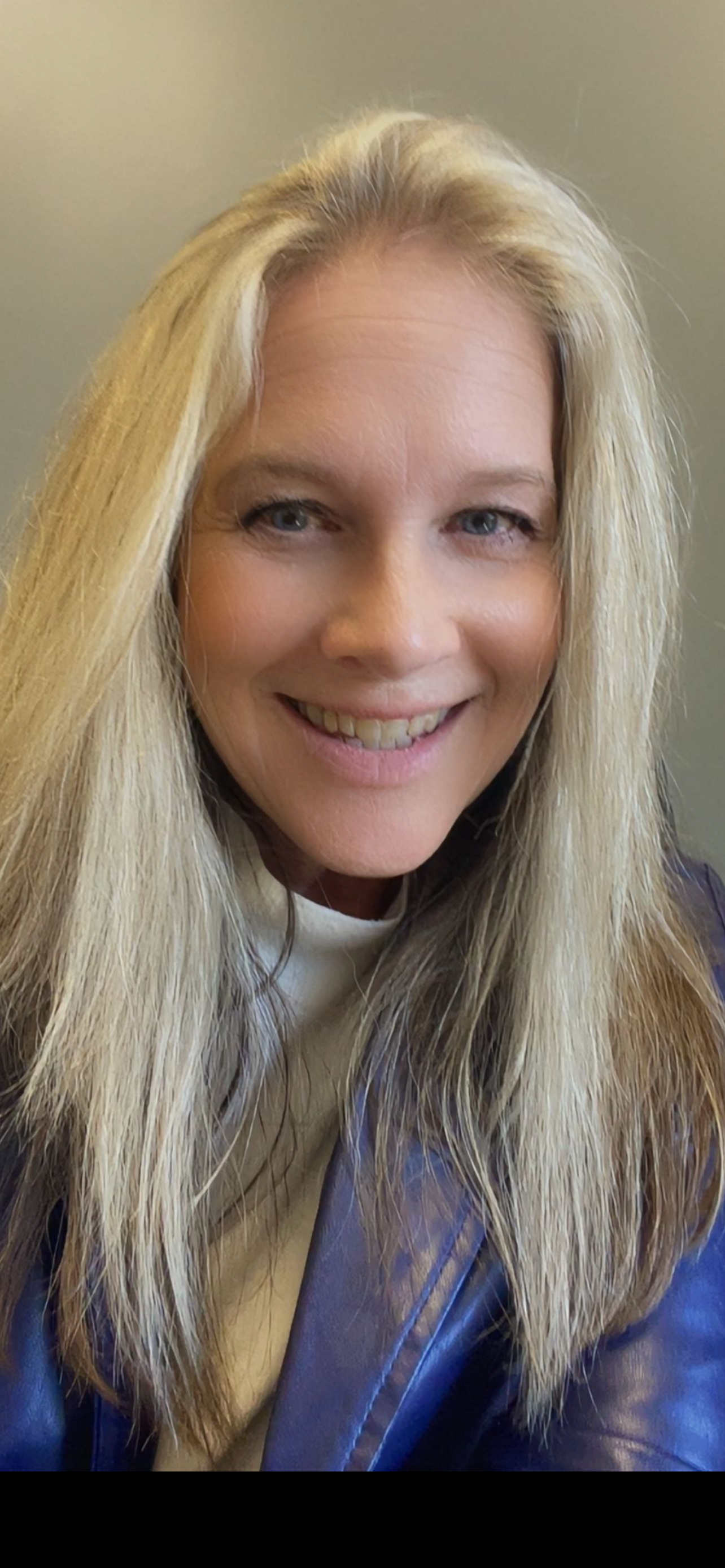 Close-up of a woman smiling with long blonde hair, wearing a blue jacket and a light-colored shirt.