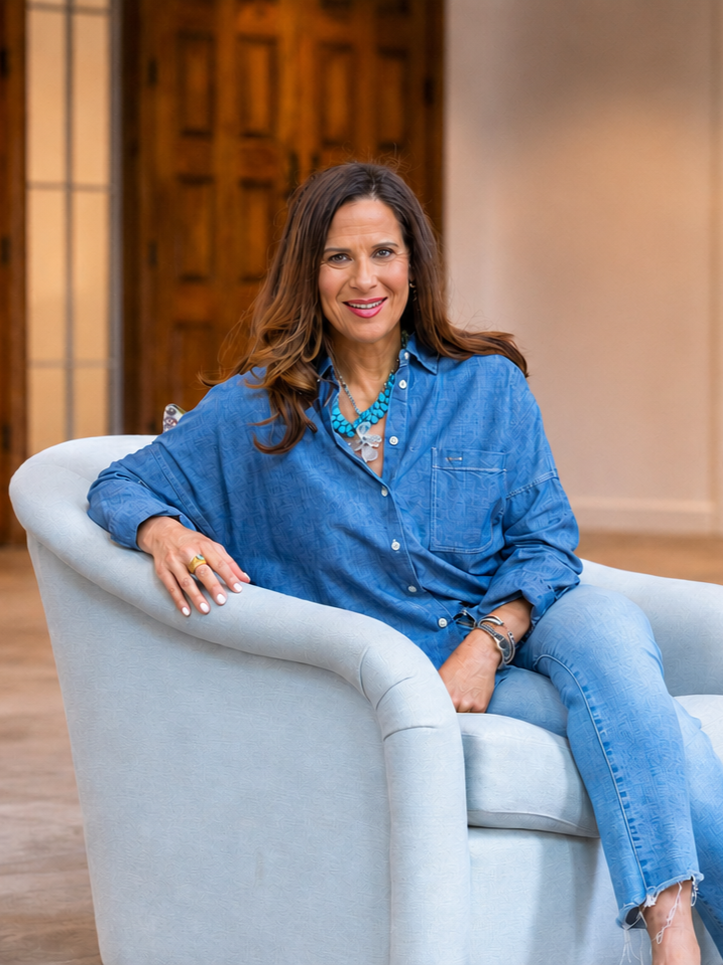 A woman with long brown hair wearing a blue denim shirt and light blue jeans sitting on a beige armchair in a cozy room with wooden doors.