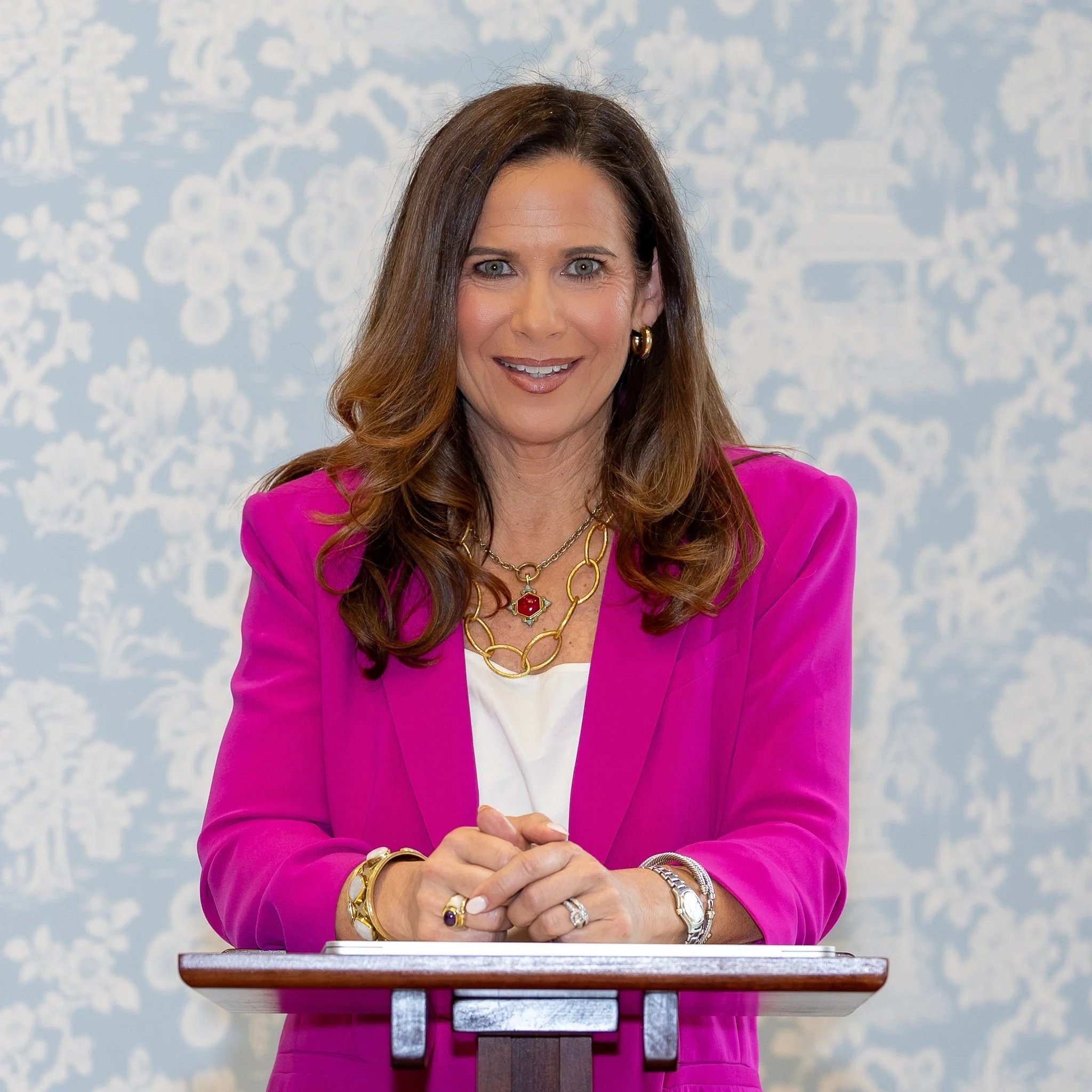 A woman with long brown hair and blue eyes, wearing a pink blazer, gold jewelry, and an expression of confidence, stands behind a wooden podium with a cloudy sky background.