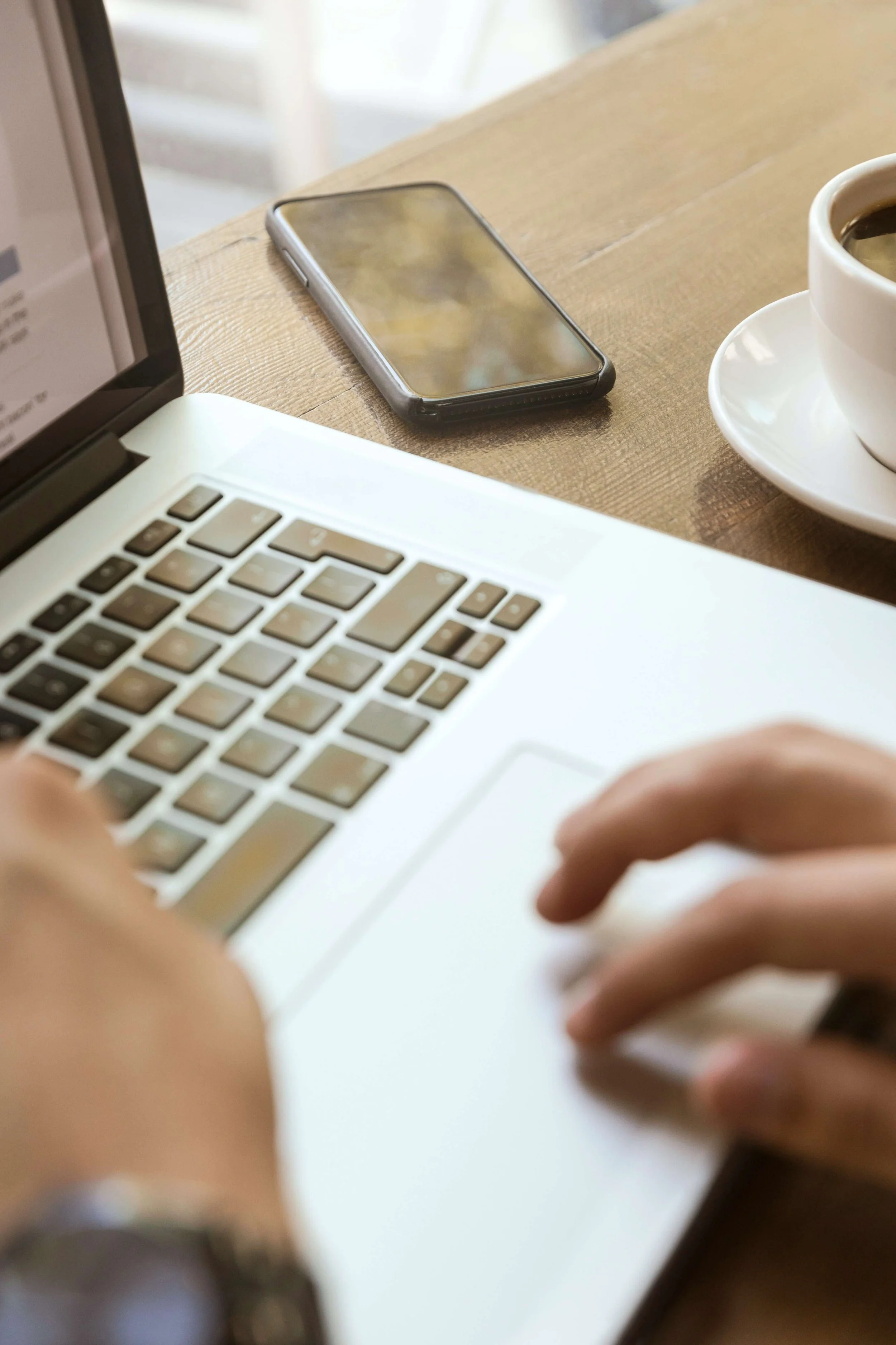 Person typing on a laptop with a smartphone and a cup of coffee on a wooden table.