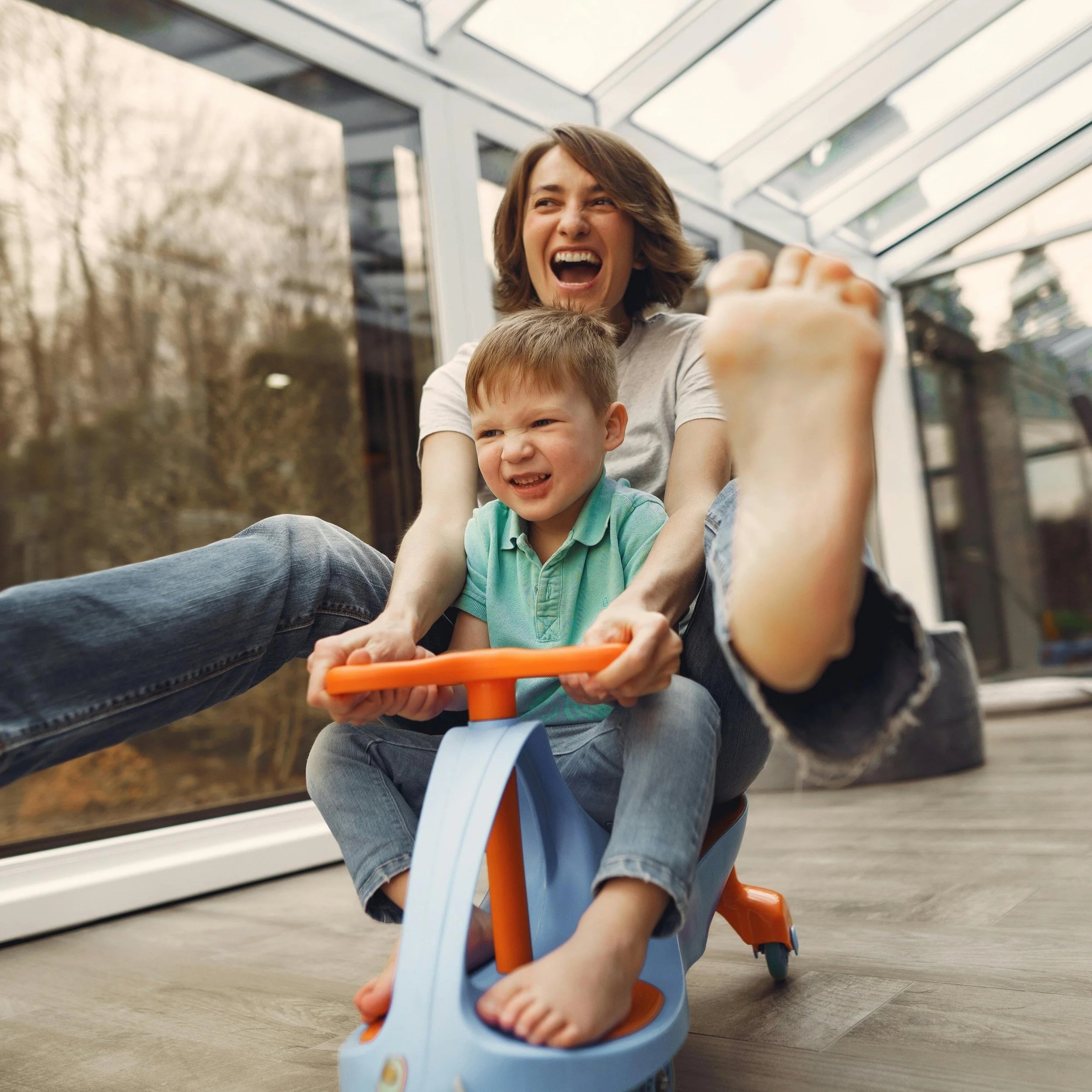 A woman and a young child riding a tricycle inside a greenhouse or glass-walled structure. The woman is smiling and making a playful face while the child is gripping the handlebars. The background shows trees and a natural outdoor setting outside the glass.