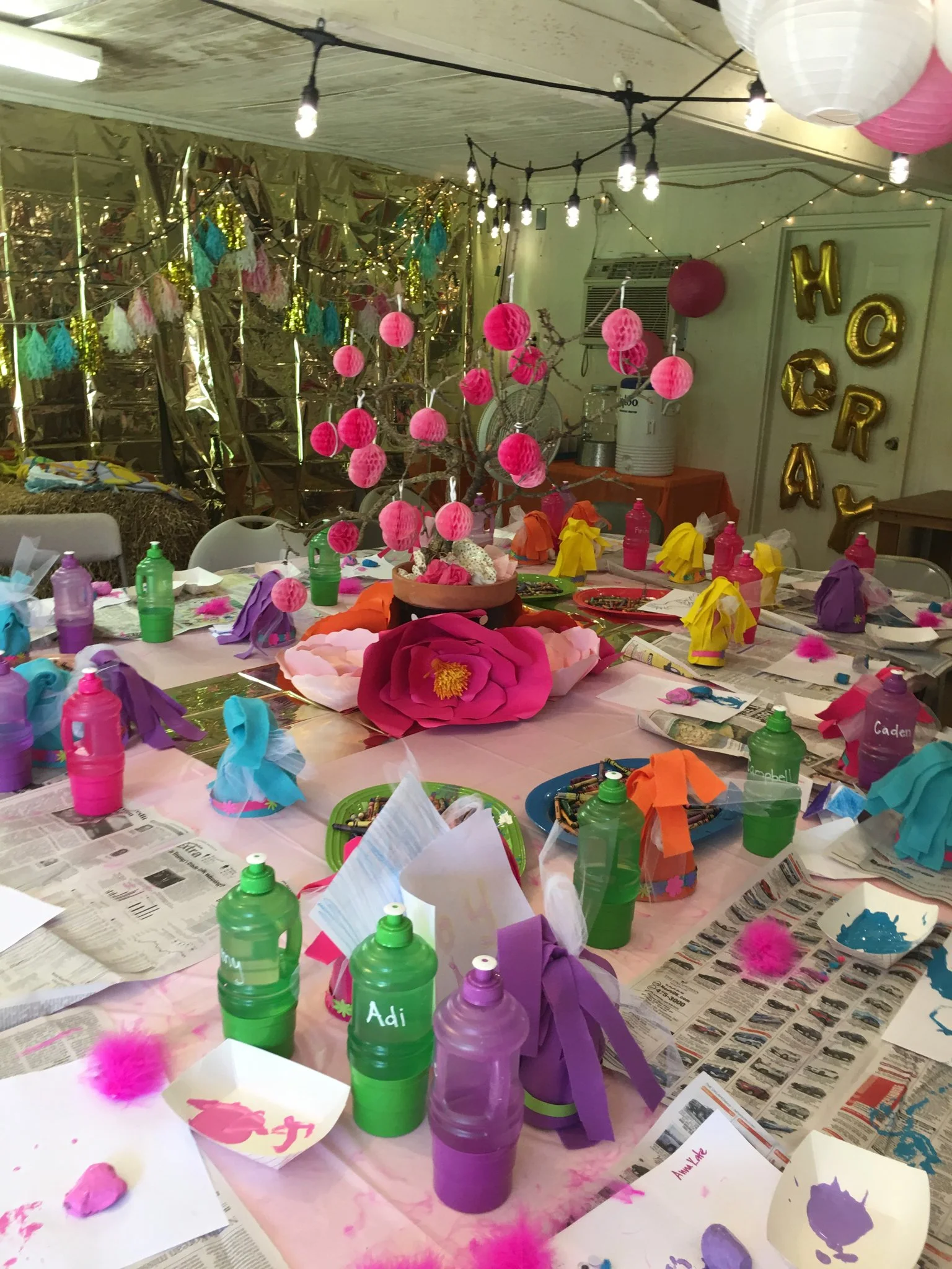 Decorative party table with colorful paper lanterns, crumpled tissue paper, and cups; background has shiny foil and string lights with hanging papel picado and balloons spelling 'H O P R A'.