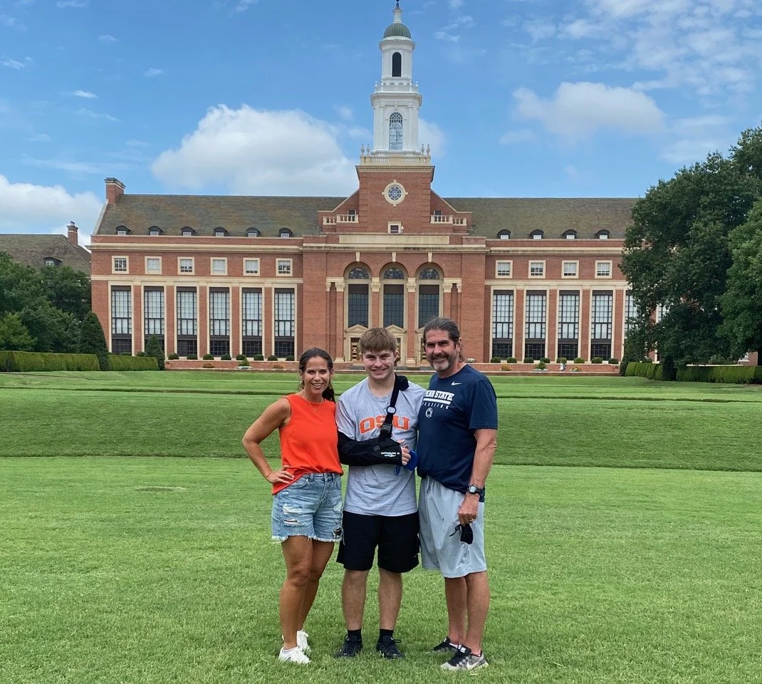 A family of three stands on a well-maintained green lawn in front of a large, historic brick building with a clock tower. The woman is wearing an orange sleeveless top and denim shorts, the young man has a gray t-shirt with orange letters and a black arm sling, and the man is in a navy shirt and gray shorts. They are smiling with an overcast sky above.