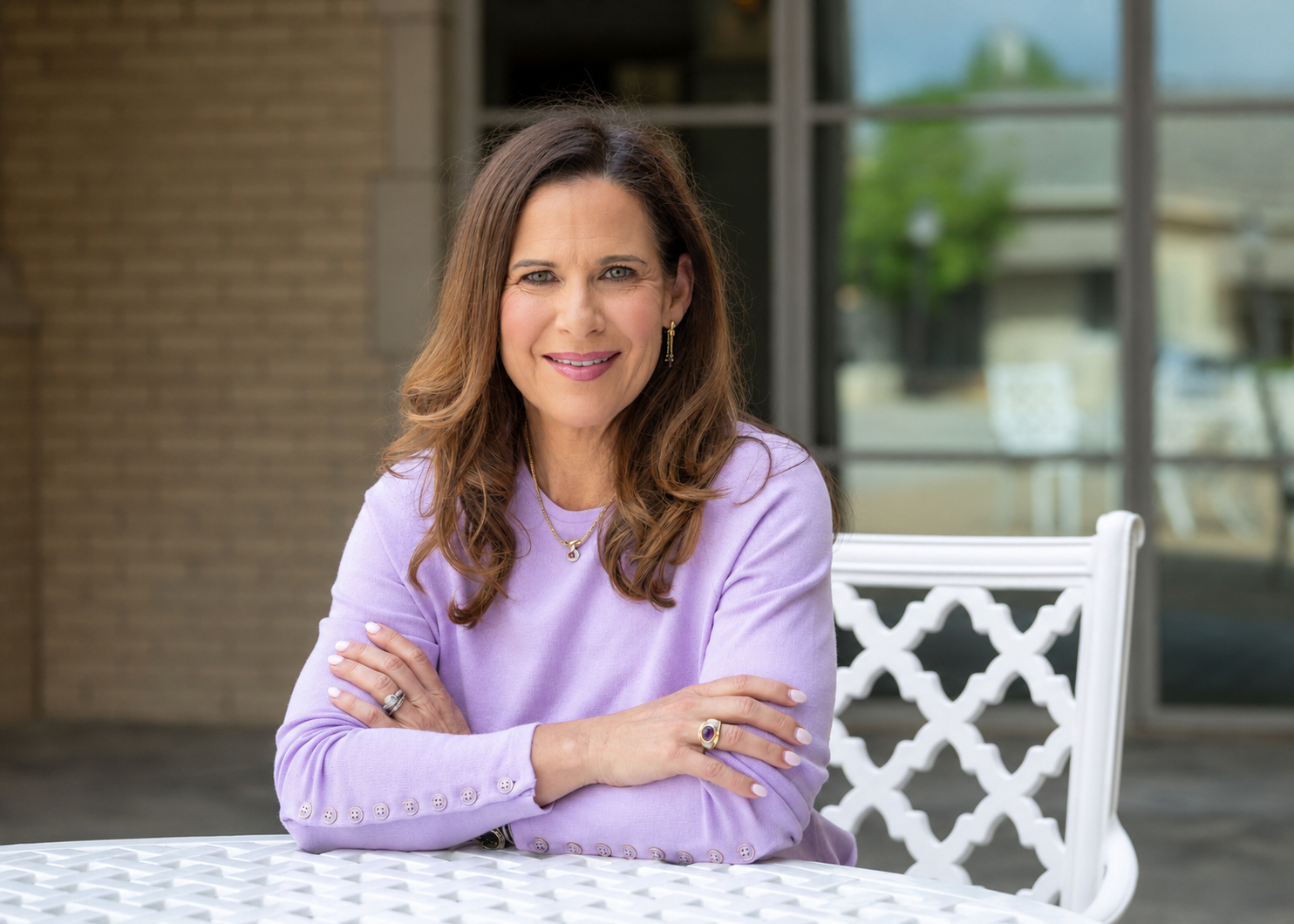 A woman with brown wavy hair, wearing a light purple sweater, gold jewelry, and rings, sitting at a white lattice table outdoors with a glass window and brick wall in the background.