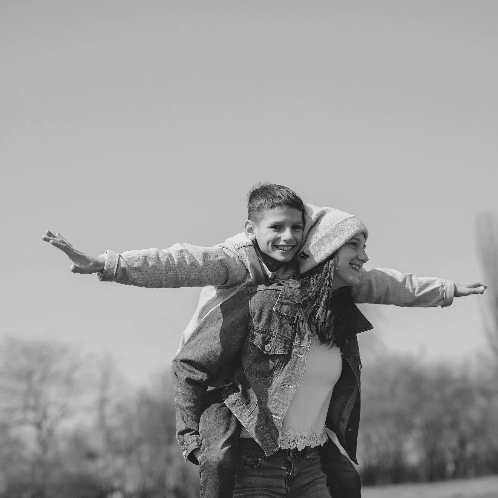 A black and white photo of a young boy smiling while riding piggyback on a woman outdoors, both with arms extended like airplane wings.