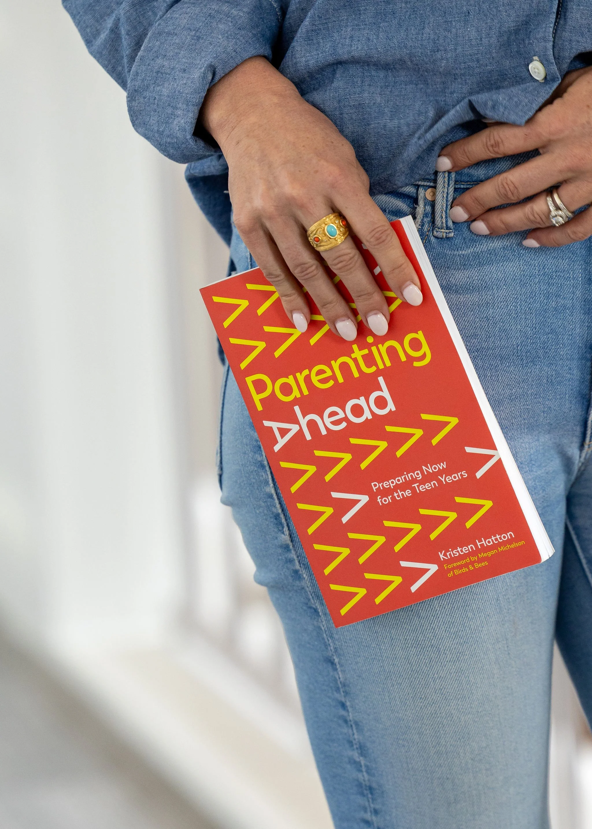 Person wearing a blue denim shirt holding a book titled "Parenting Ahead" with a red cover and yellow arrow designs, with their hand on their hip and a gold ring with a turquoise stone on their finger.