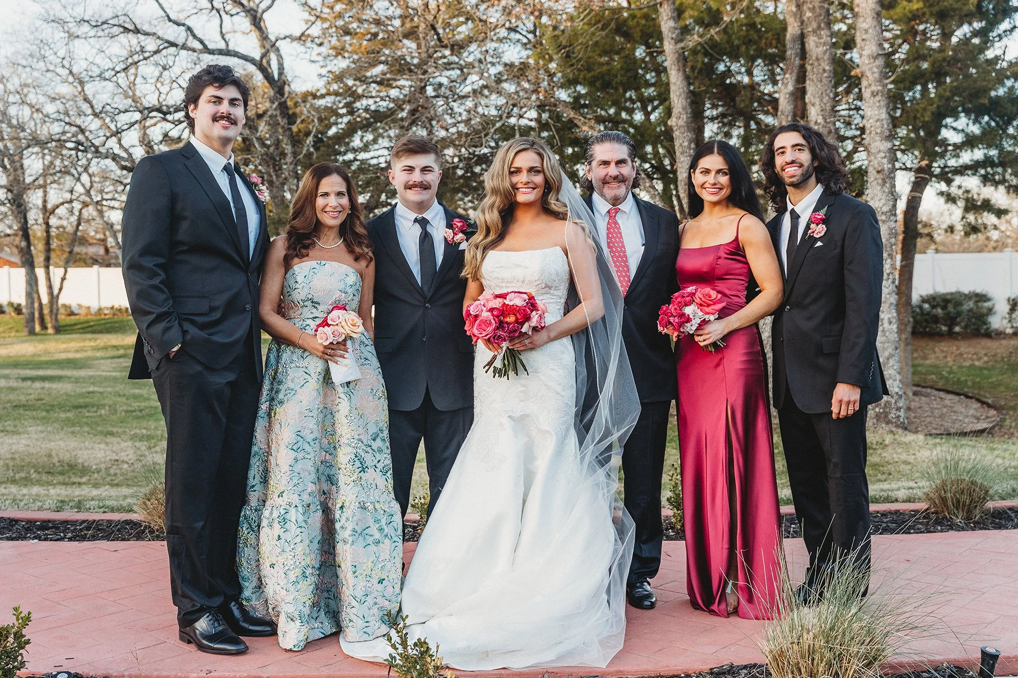 Group of seven people at a wedding outdoors during fall, standing on a pathway with trees and a white fence in the background. The bride, in a white wedding dress holding a bouquet, is in the center. Two men and four women, dressed in formal attire, are positioned around her, with the men in suits and women in colorful dresses. Everyone is smiling.